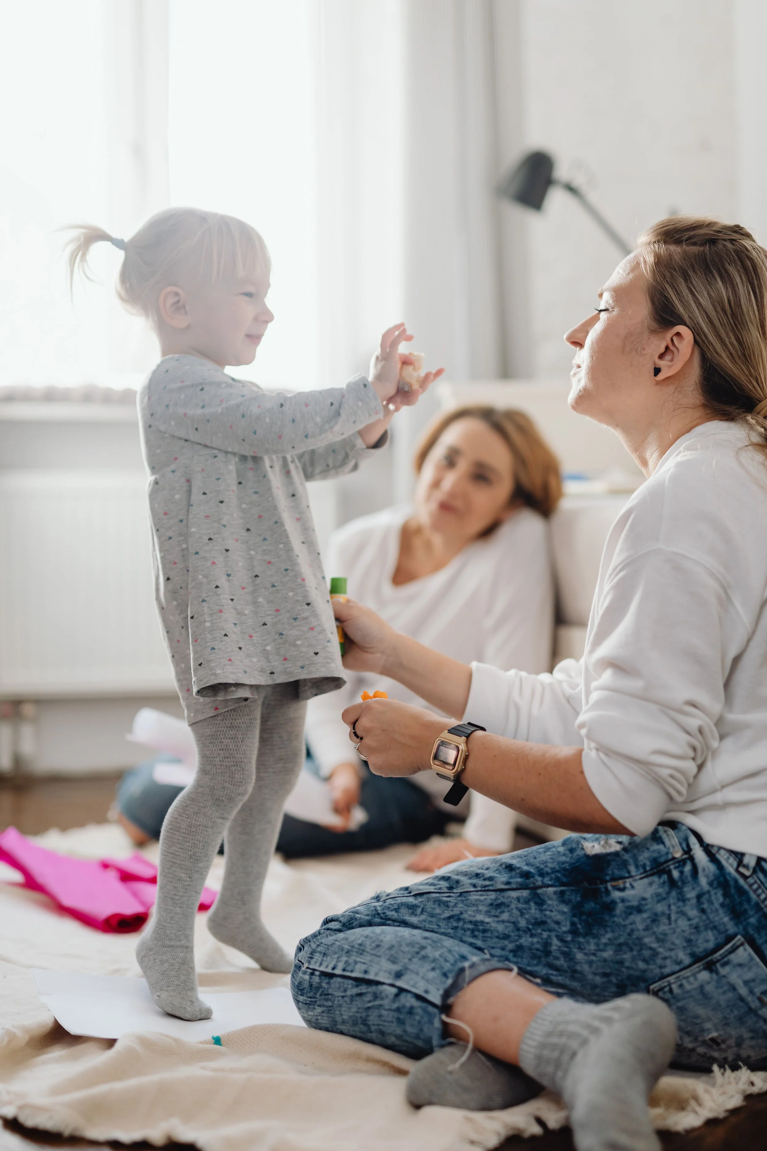 A young girl with blonde hair tied in a ponytail is standing and smiling at an adult woman. The woman is kneeling on the floor, wearing a white shirt and denim jeans, and is handing something to the girl. An older woman with red hair is sitting in the background, observing the interaction. The scene takes place in a bright room with natural light coming through sheer curtains.