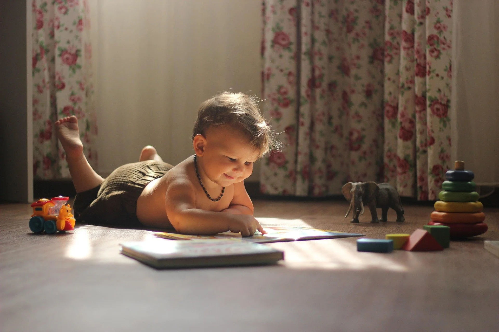 Young boy lying on the floor, playing with a book, with toys around him in a room with floral curtains.