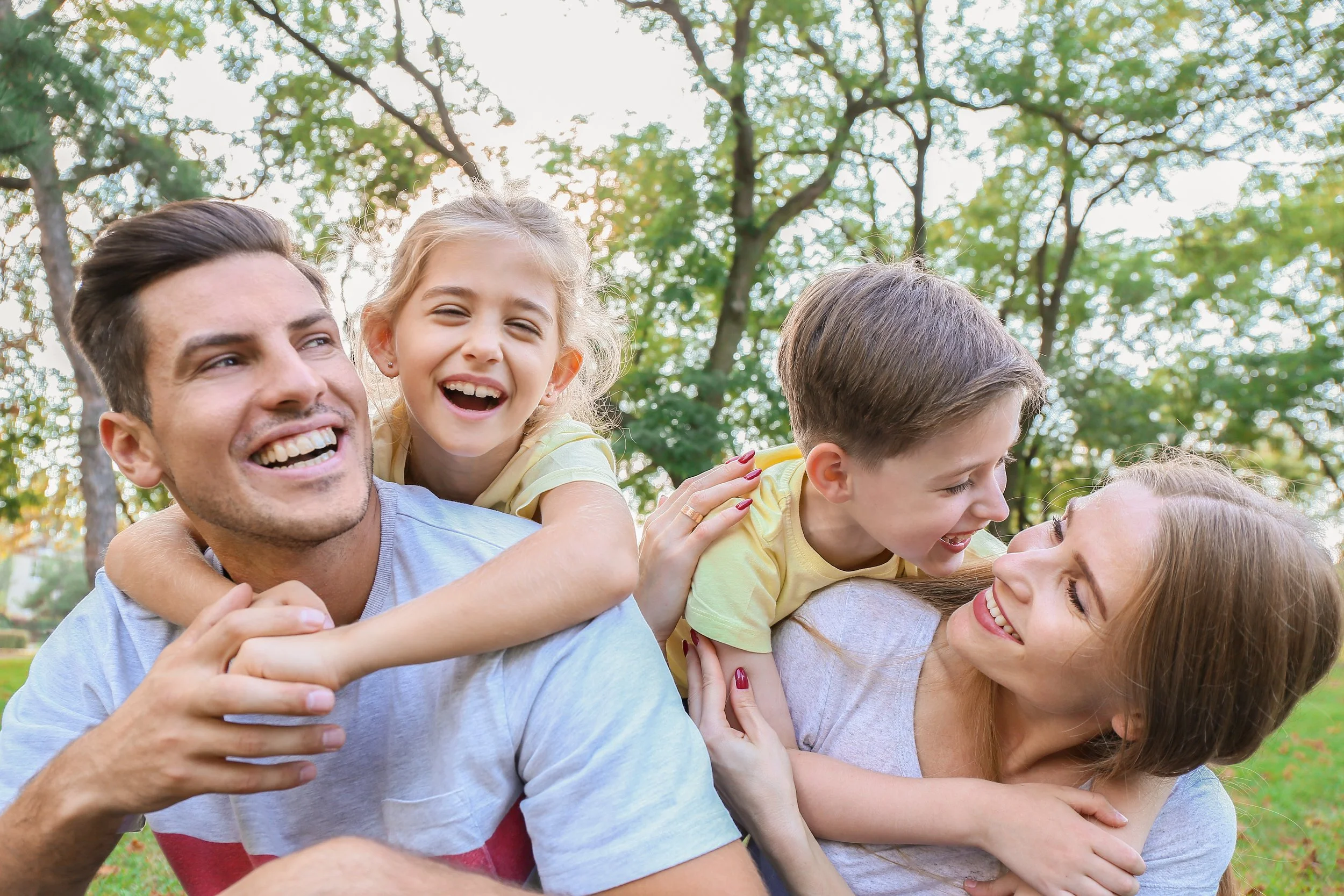 A man giving a piggyback ride to a smiling young girl while other children and woman are playing and laughing outdoors in a park with trees in the background.