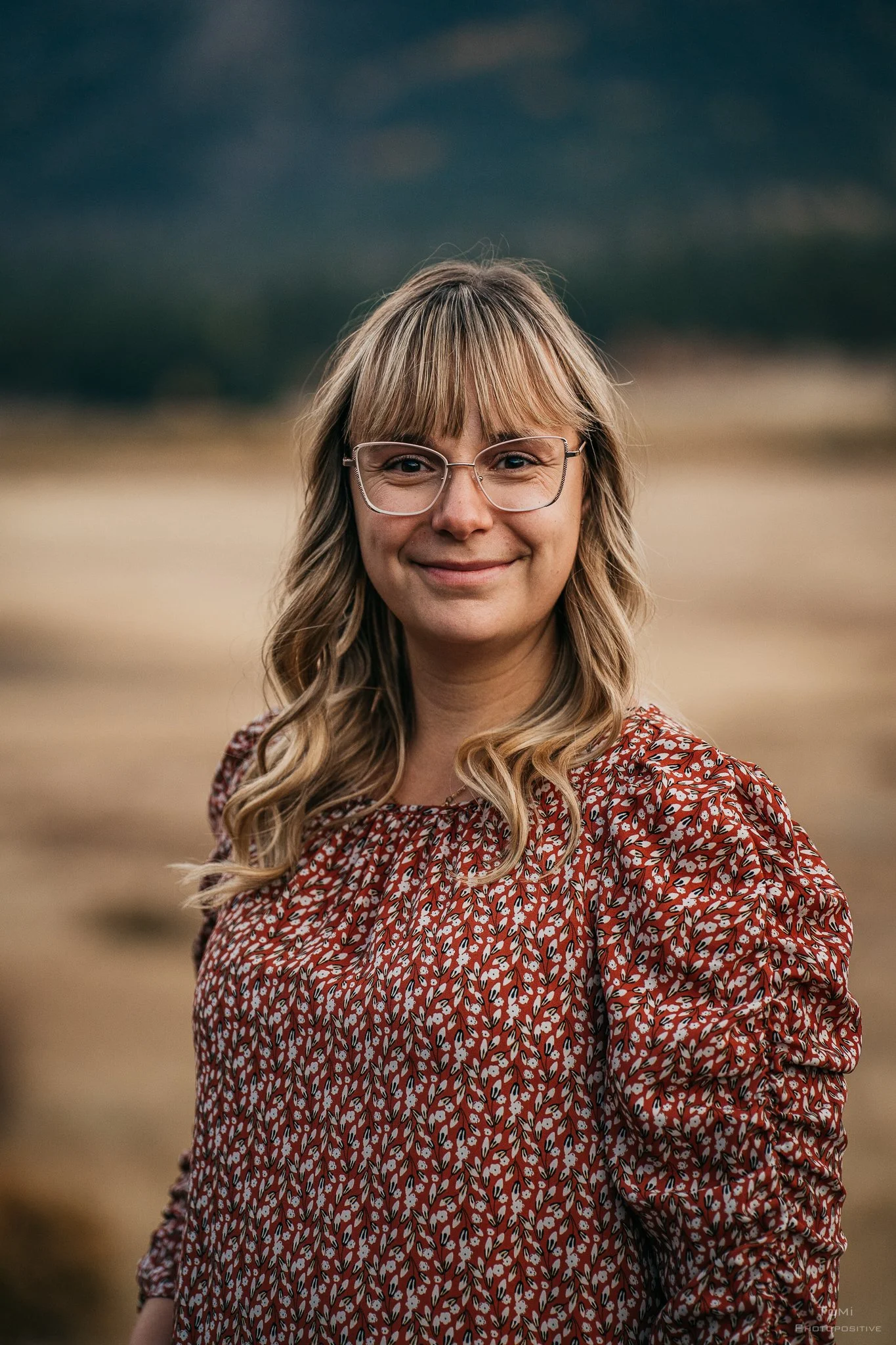 A friendly woman with wavy blonde hair and glasses smiling outdoors, wearing an orange patterned blouse, with a blurred landscape in the background.
