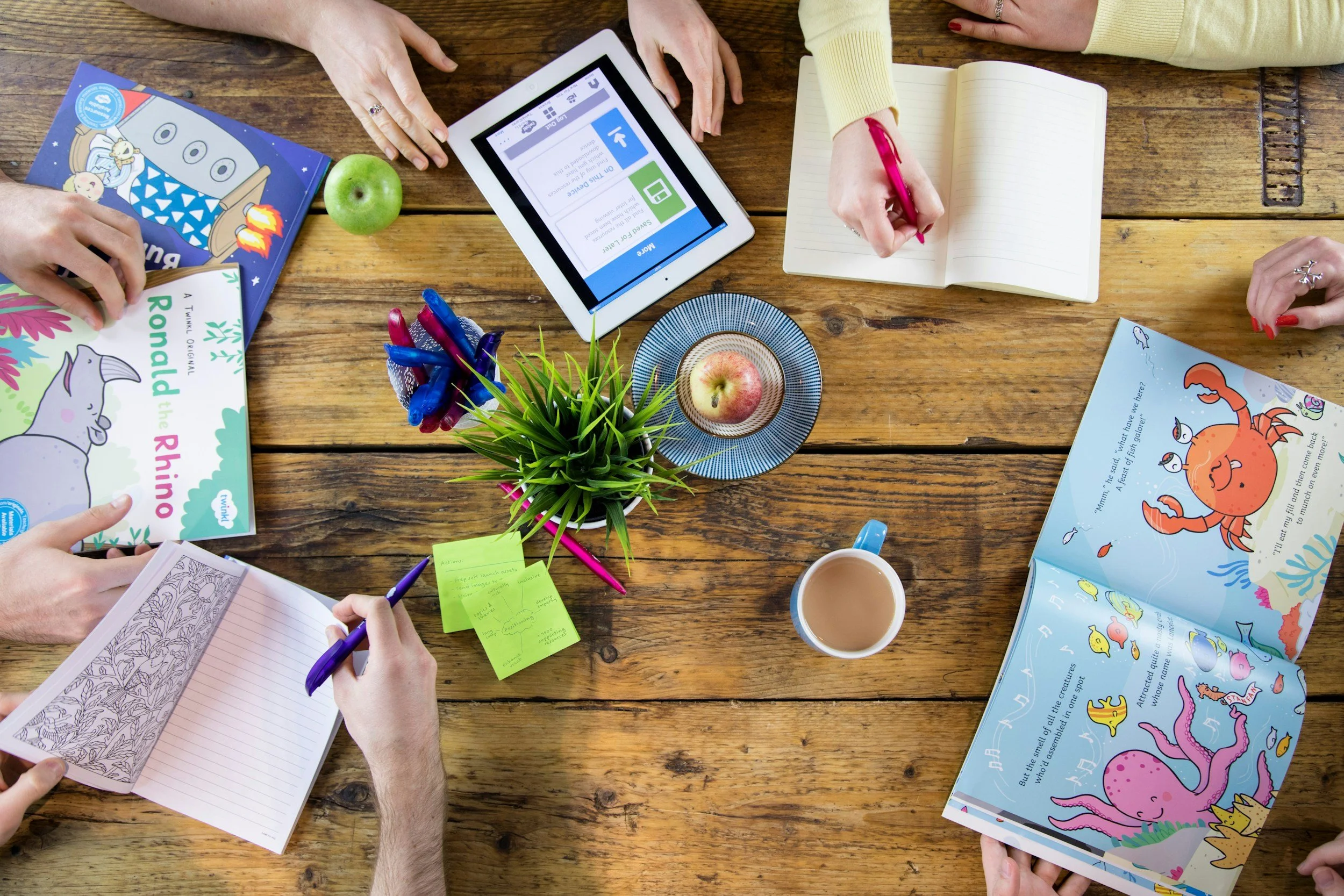 Multiple people are gathered around a wooden table working on books, a tablet, notebooks, and colorful pens, with a green apple, a small plant, and a coffee mug also on the table.