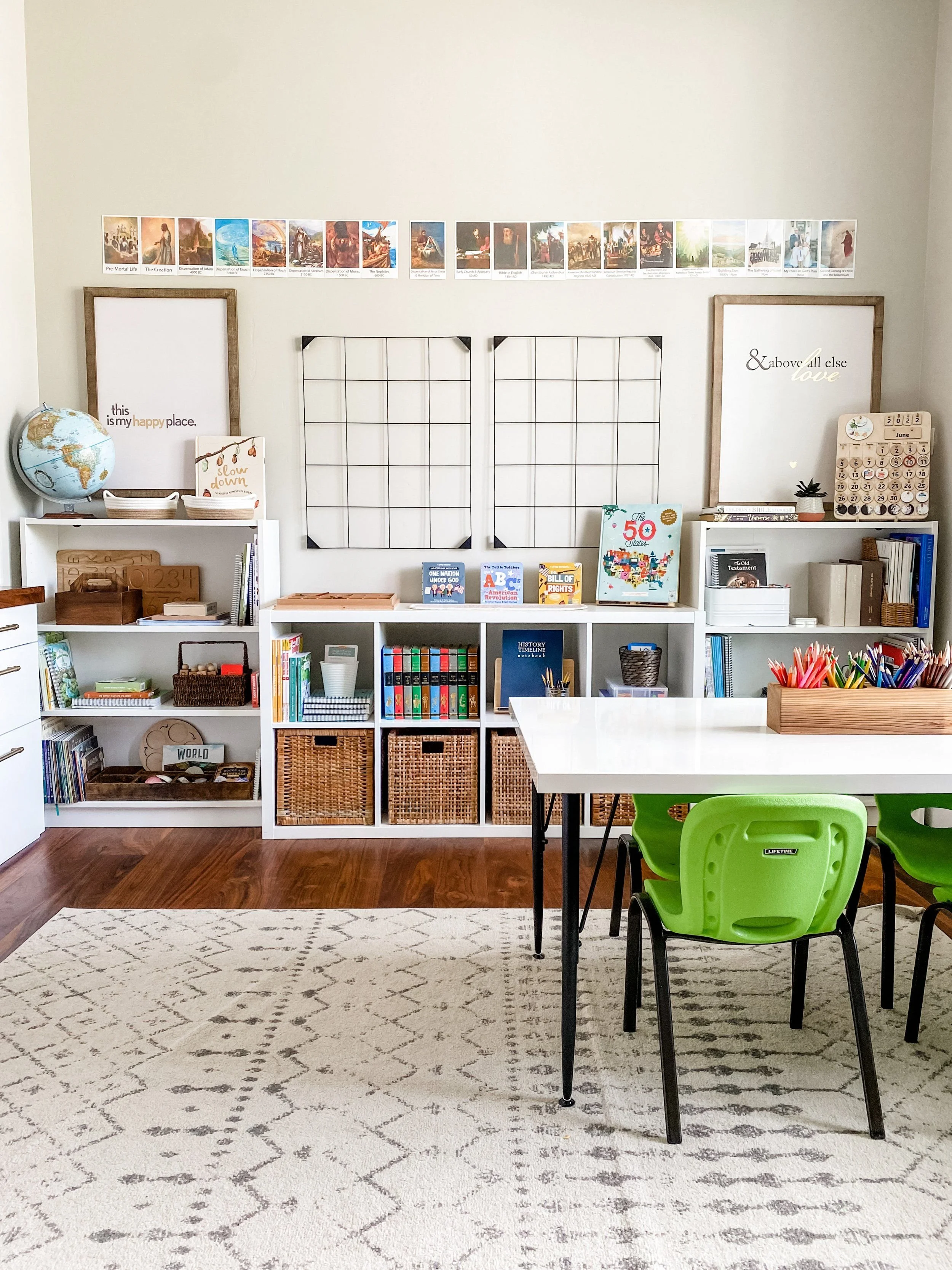 A bright and organized classroom with white bookshelves, a white table with green chairs, framed wall art, educational books, a globe, and decorative items, with a patterned white and black rug on the wooden floor.