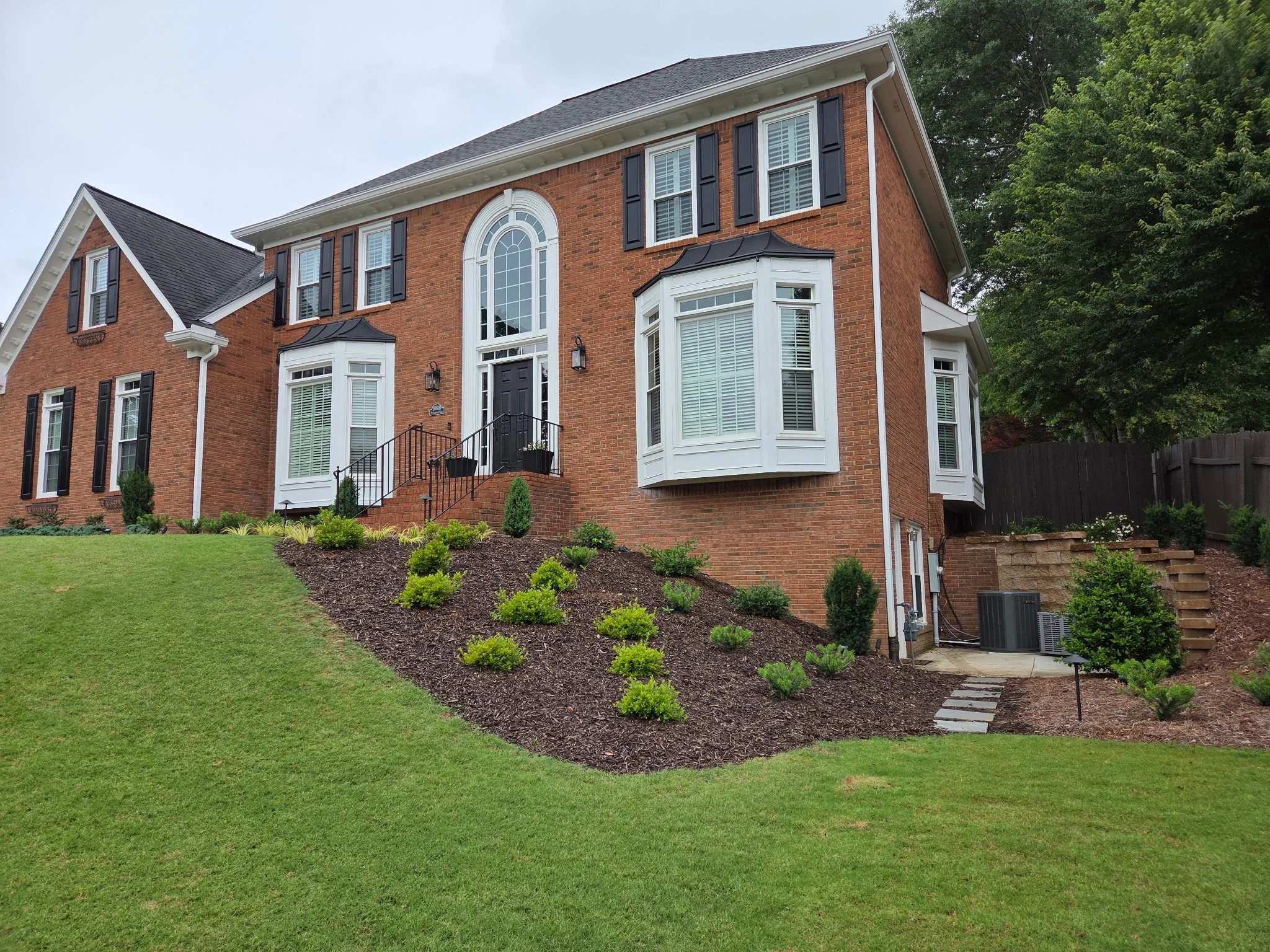 A large brick house with white trim, black shutters, and a black front door, surrounded by a well-maintained lawn and landscaping. There are trees and a wooden fence in the background.