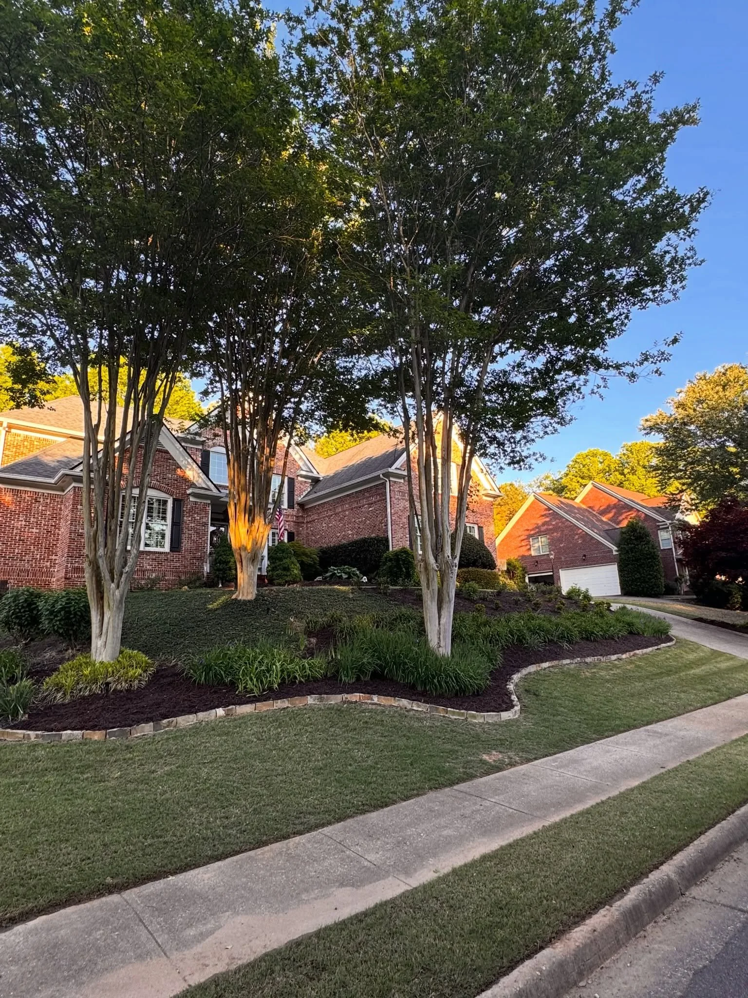 A suburban neighborhood with trees, brick houses, manicured lawns, and a sidewalk in the foreground. The scene is illuminated by streetlights as evening approaches.