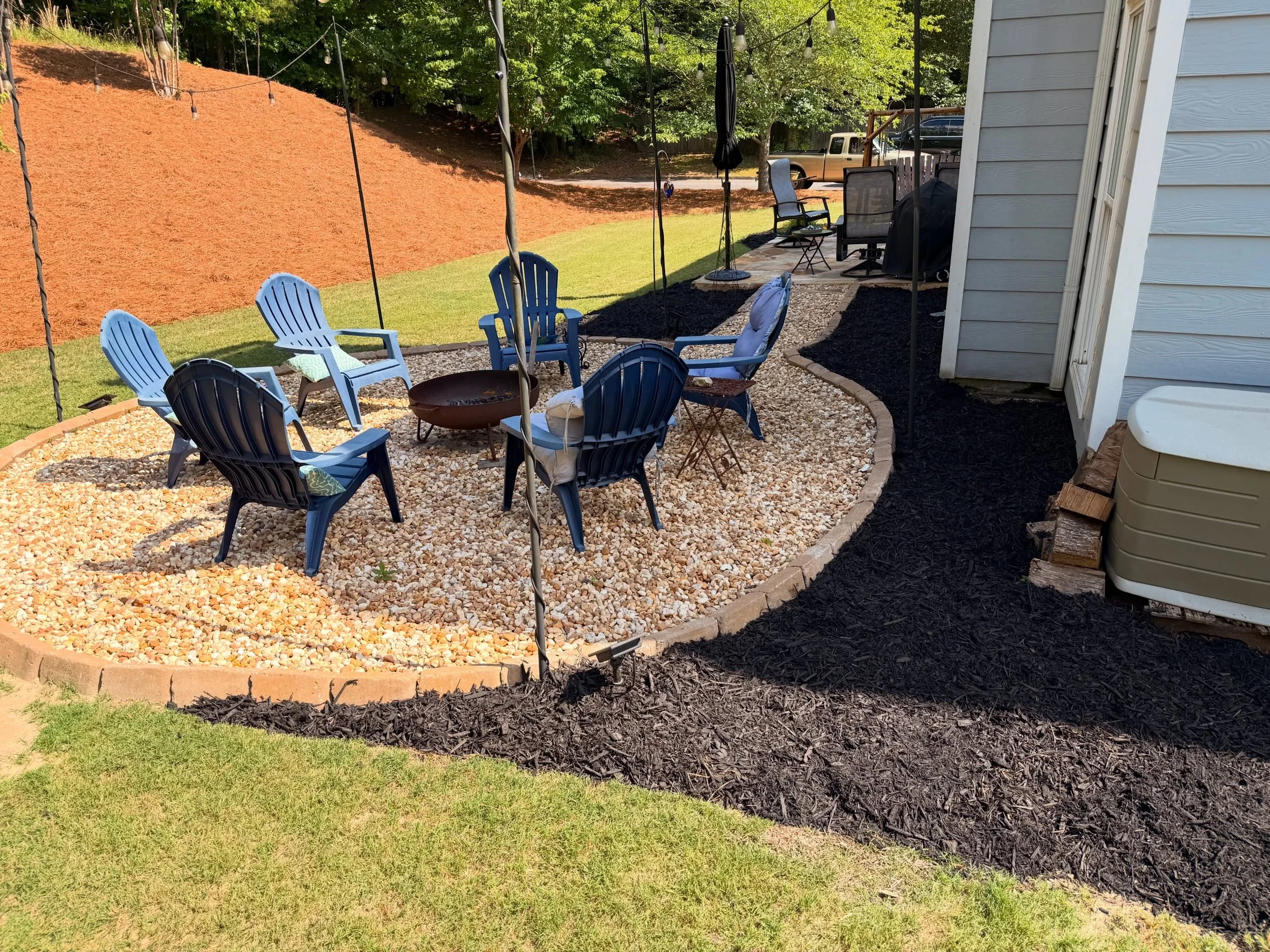 Backyard patio with blue chairs, a firepit, and a stone path, surrounded by a grassy yard, mulched borders, and a retaining wall with trees in the background.