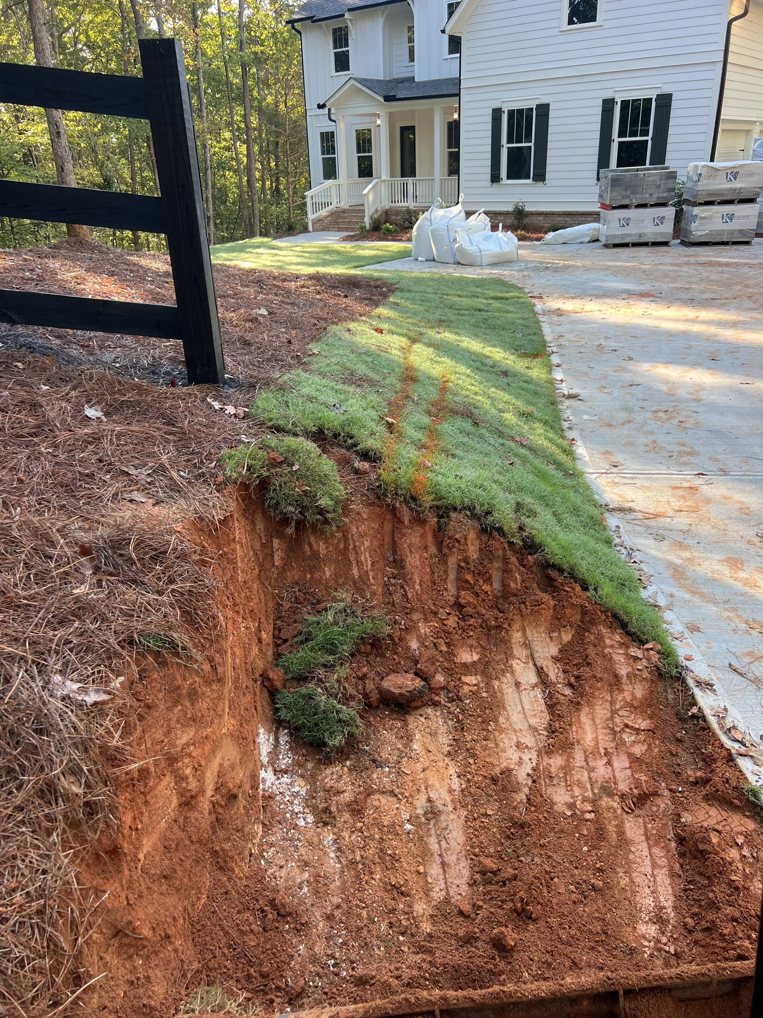 A deep trench dug in front of a white house with black shutters, with construction materials such as bags of dirt and bricks nearby. The yard is partially grassy with trees in the background.