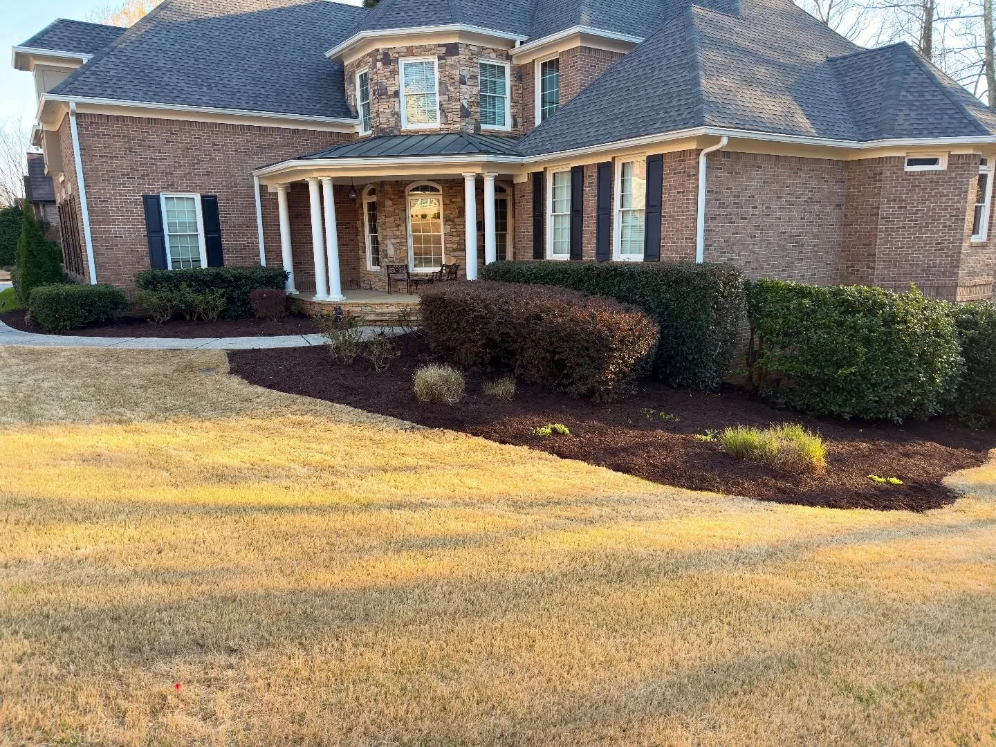 Front view of a large brick house with a curved porch supported by white columns, surrounded by landscaped bushes and mulch, with a well-maintained lawn.
