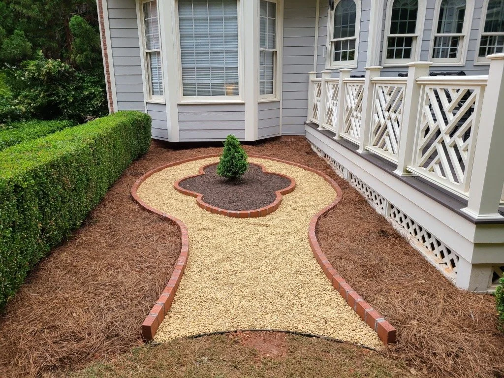 A small landscaped backyard area with a curved gravel pathway bordered by bricks, leading to a small shrub in a circular brick bed, adjacent to a light-colored house with a porch and white decorative railing.