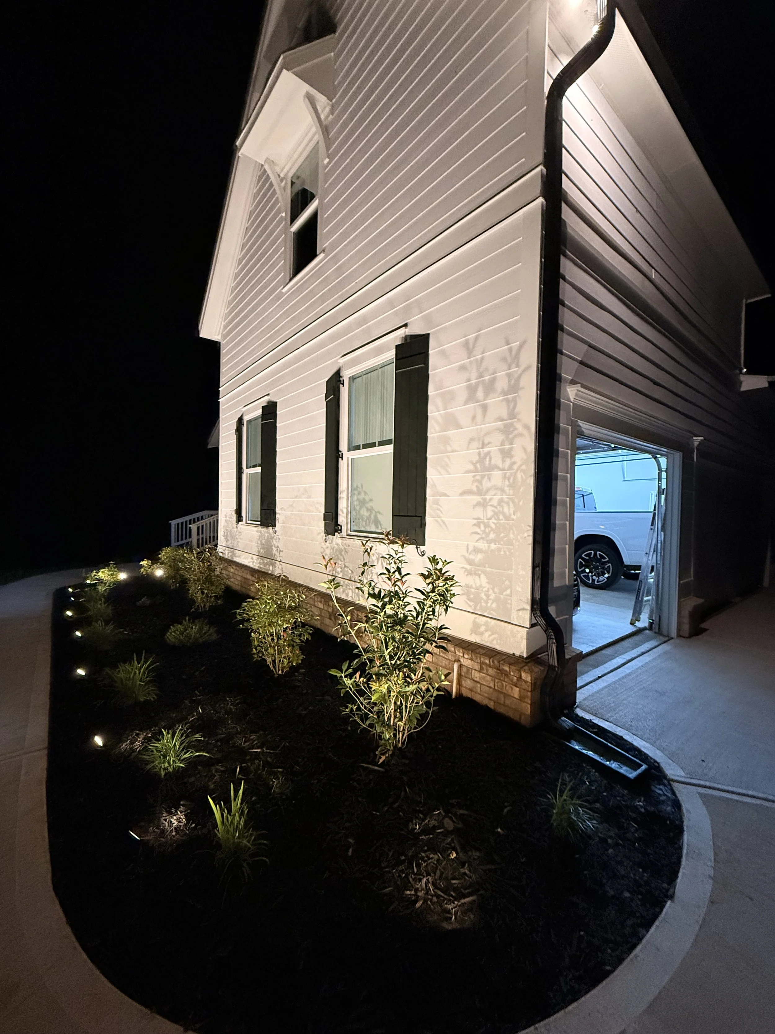 Nighttime exterior view of the side of a white house with a garage. The house has two windows with black shutters. The garden bed with small plants and landscape lights is in the foreground. The garage door is open, revealing a white vehicle parked i