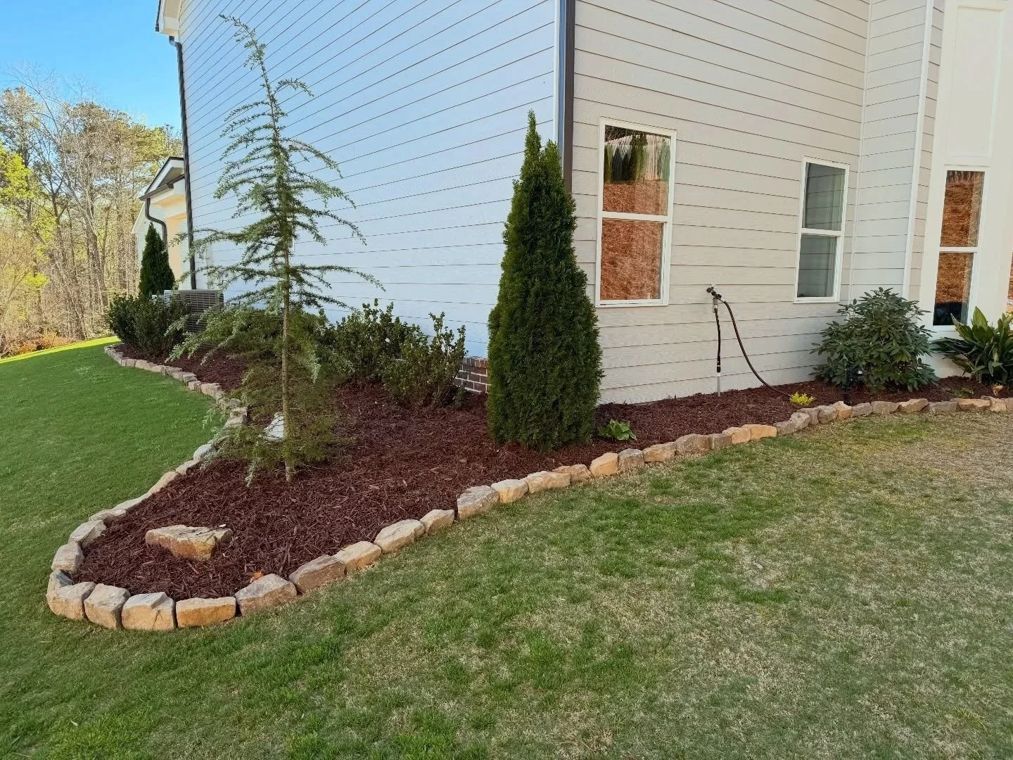 A landscaped yard with a flower bed bordered by stones, containing various bushes and a small tree, next to a white house with three windows and an outdoor faucet with a hose. The background shows a green lawn and a wooded area.