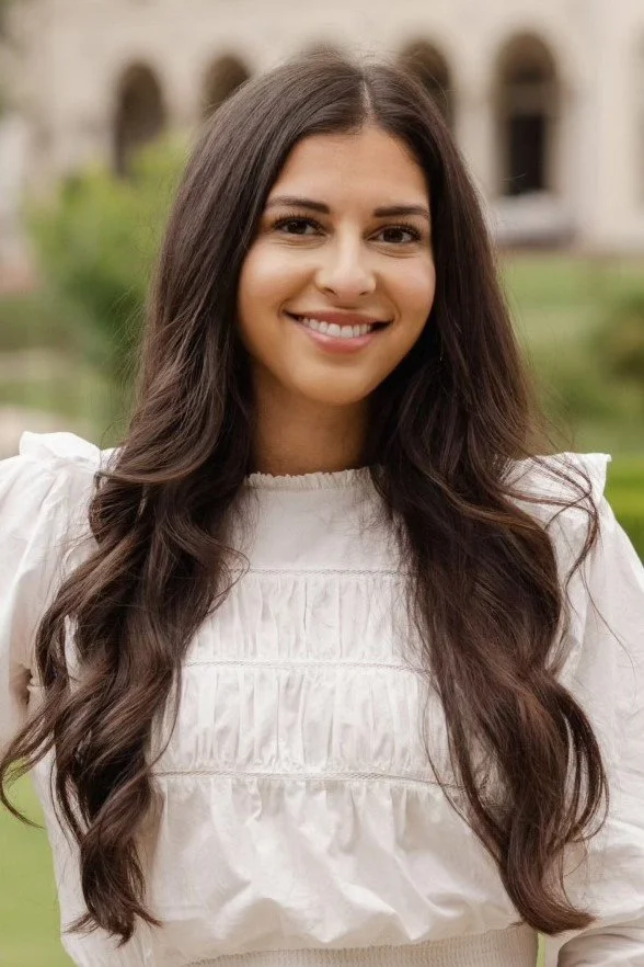 A woman with long, wavy brown hair smiling outdoors in front of a blurred background with a building and greenery.