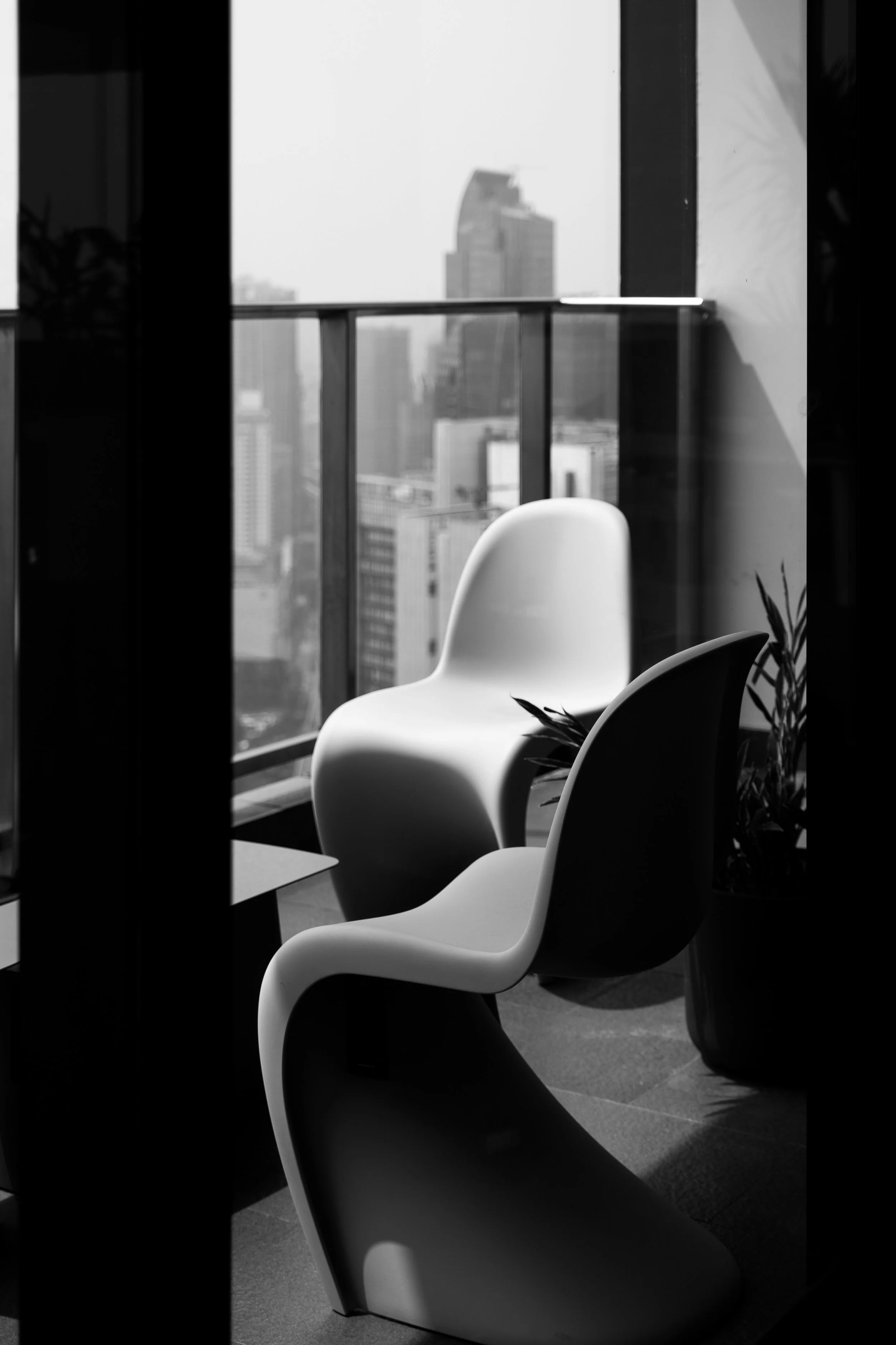 Black and white photo of a city view from an office window. Two modern chairs are visible inside the office, with a plant on the right. The city skyline features tall skyscrapers, one with a distinctive sloped roof, outside the large window.
