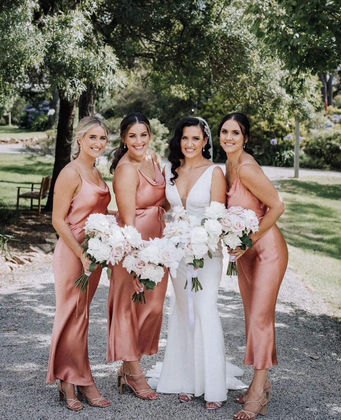A bride in a white wedding dress and four bridesmaids in pink satin dresses holding bouquets of white roses outdoors in a garden setting.