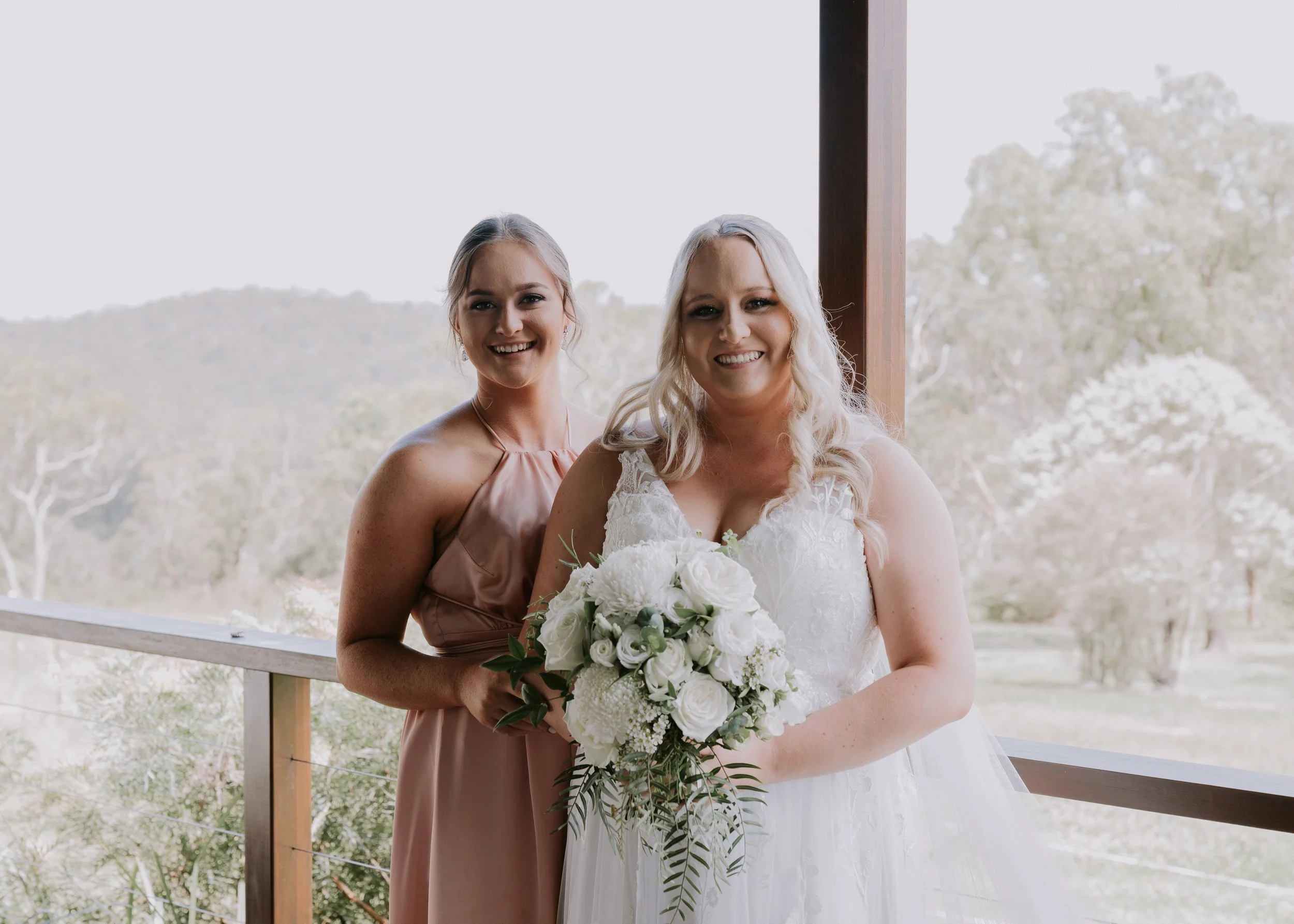 Two women, one in a wedding dress holding a bouquet, and the other in a mauve dress, standing together outdoors on a porch with trees in the background.