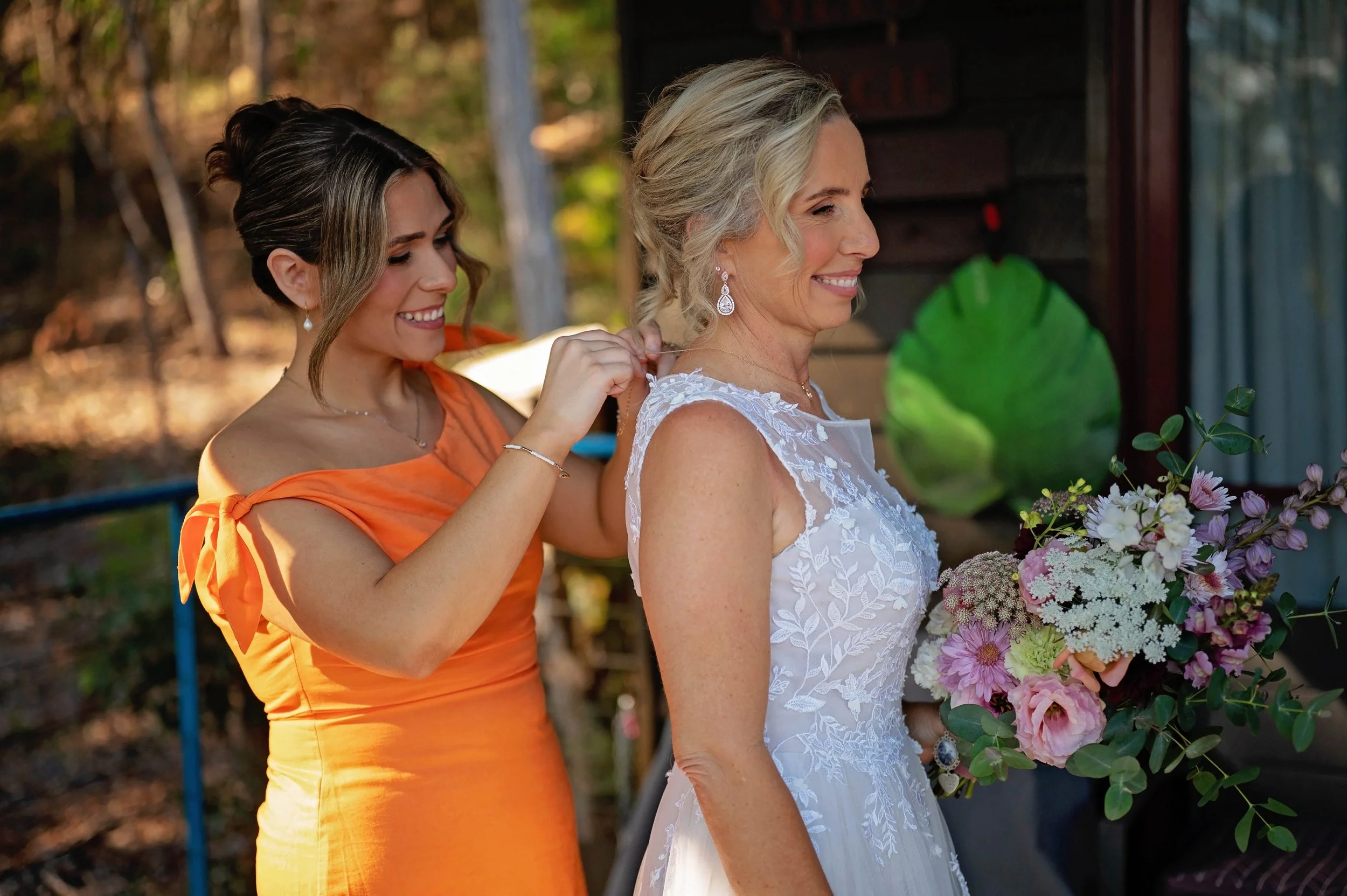 A woman in a white dress holding a bouquet of pink and white flowers stands outdoors, smiling, as another woman in an orange dress helps her with her necklace. They are near a green plant with a blurred background of trees.