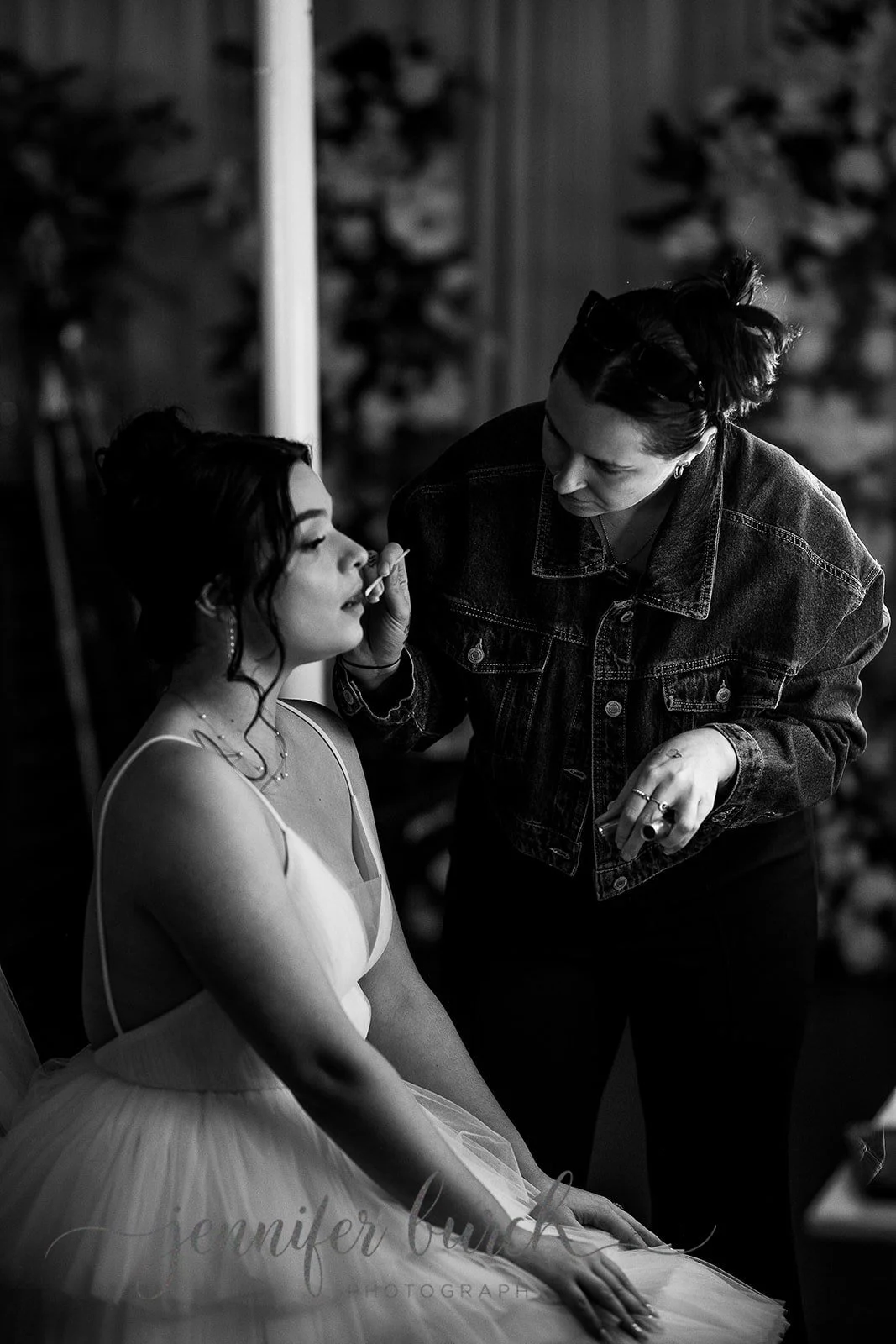 A makeup artist applying lipstick to a young woman in a white dress, sitting with her eyes closed, during a photo shoot.