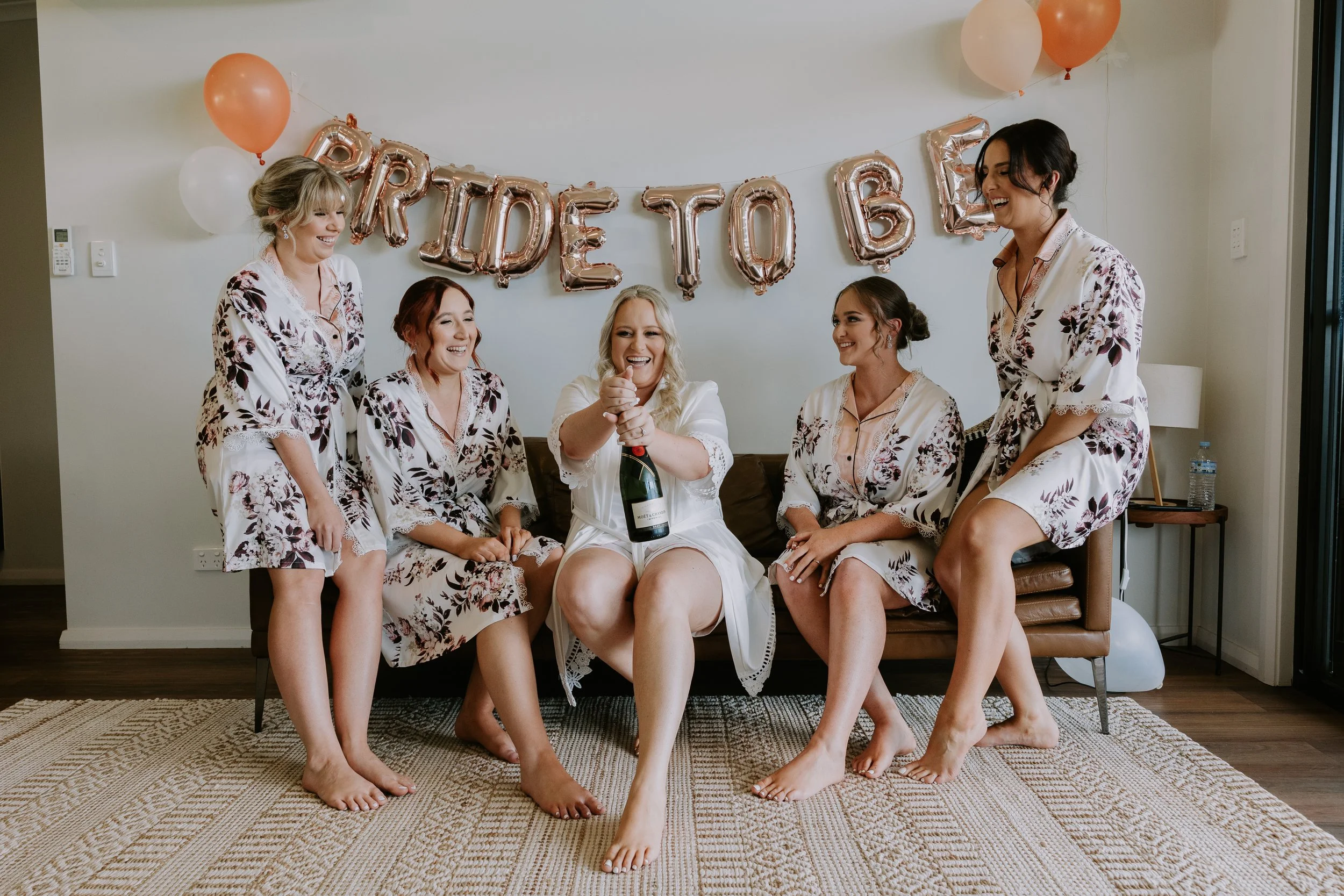 A group of five women in pajamas celebrating a birthday with balloons and shiny letter balloons spelling 'HAPPY BIRTHDAY' in the background. One woman is opening a bottle of champagne while all are smiling and sitting on a couch in a living room.