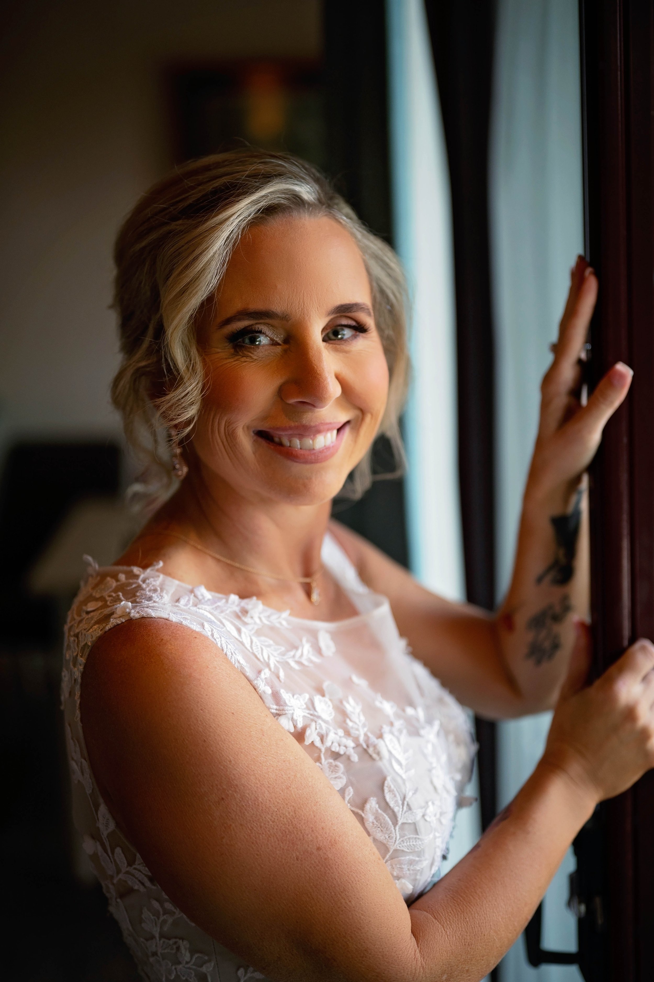 A smiling bride with short blonde hair, wearing a white lace dress, is looking at the camera and holding onto a dark wooden door frame or window ledge.