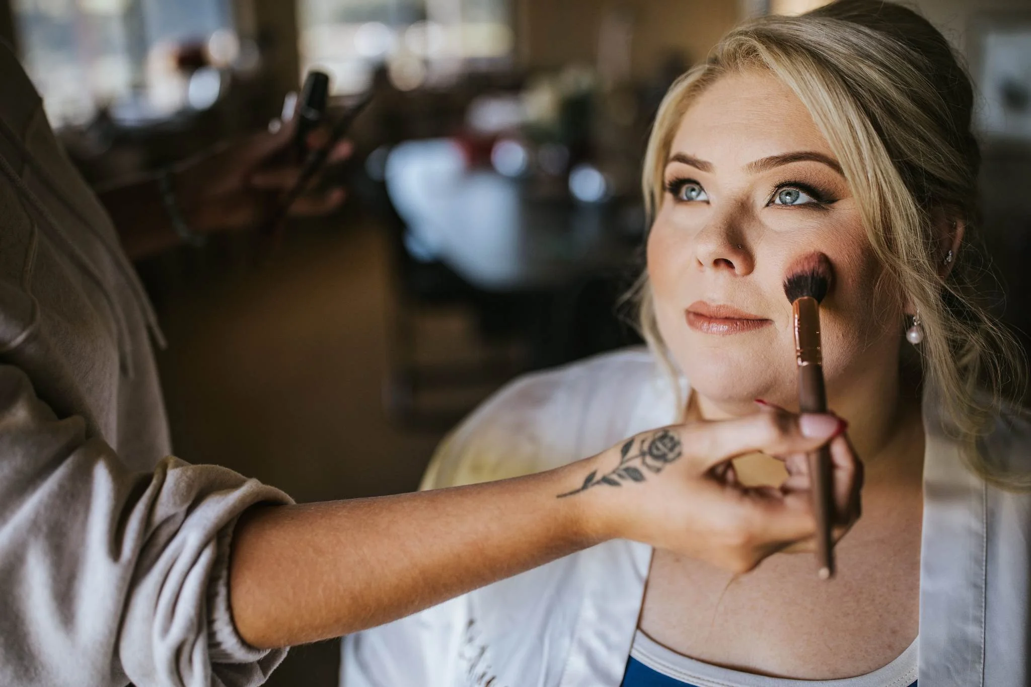 A woman with blonde hair and blue eyes getting makeup applied to her face by a person with a tattooed arm. The woman is looking slightly upwards with a calm expression.
