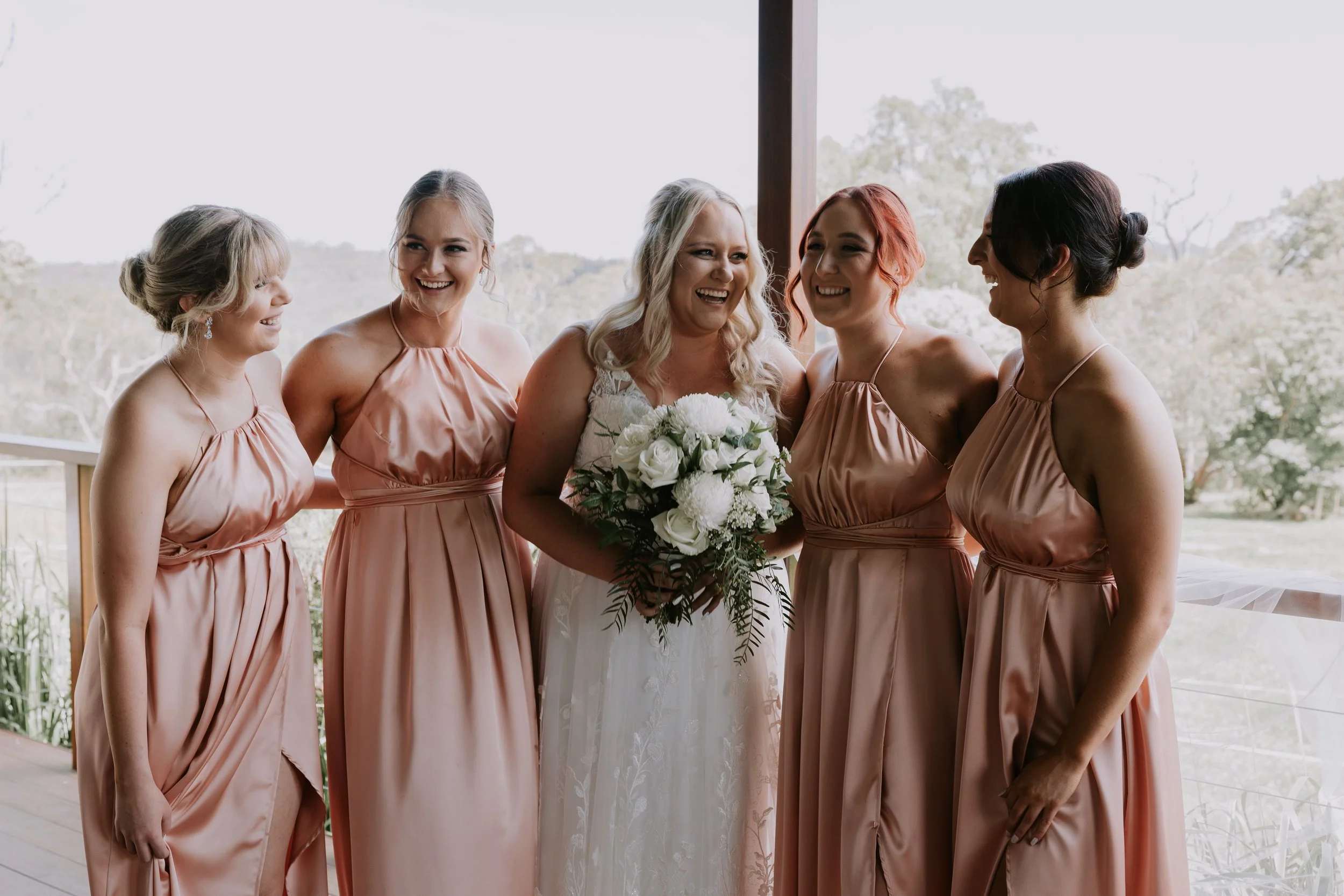 A bride and four bridesmaids standing together outside, smiling and sharing a happy moment. The bride is holding a bouquet of white flowers and wearing a white wedding dress. The bridesmaids are dressed in matching peach-colored dresses.