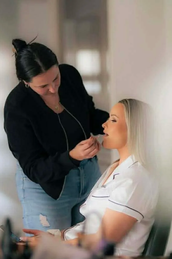 A woman in a white pajama top sits with her eyes closed as another woman applies makeup or lipstick to her lips in a room with neutral-colored walls.