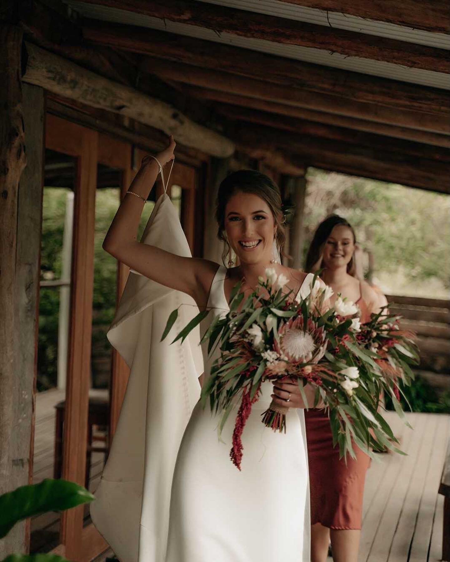 A bride in a white wedding dress holding a bouquet of flowers, smiling, with another woman in the background on a wooden porch.