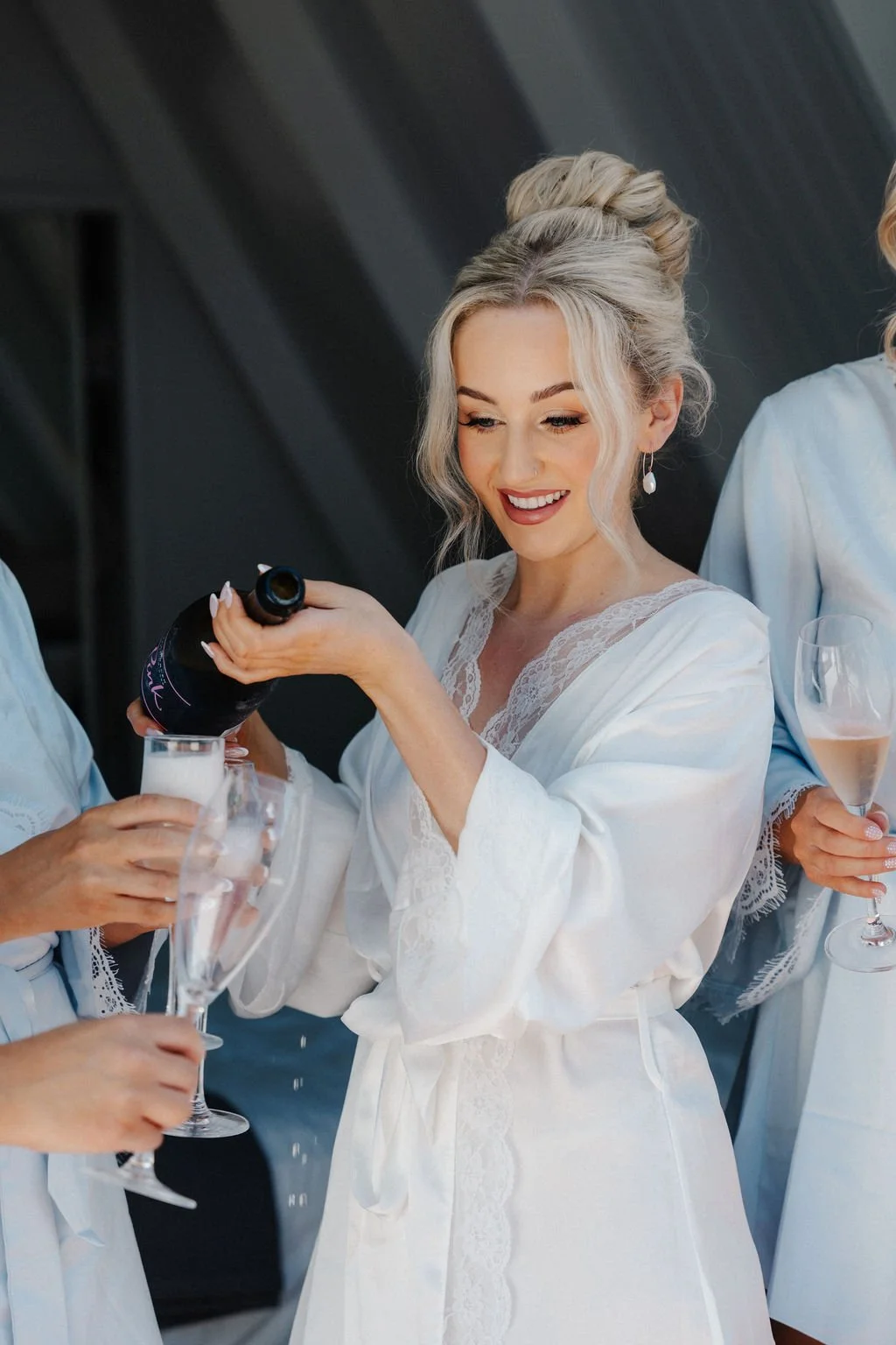 A bride in a white satin robe with lace trim is pouring champagne into a glass at a wedding celebration, smiling as she prepares to toast.