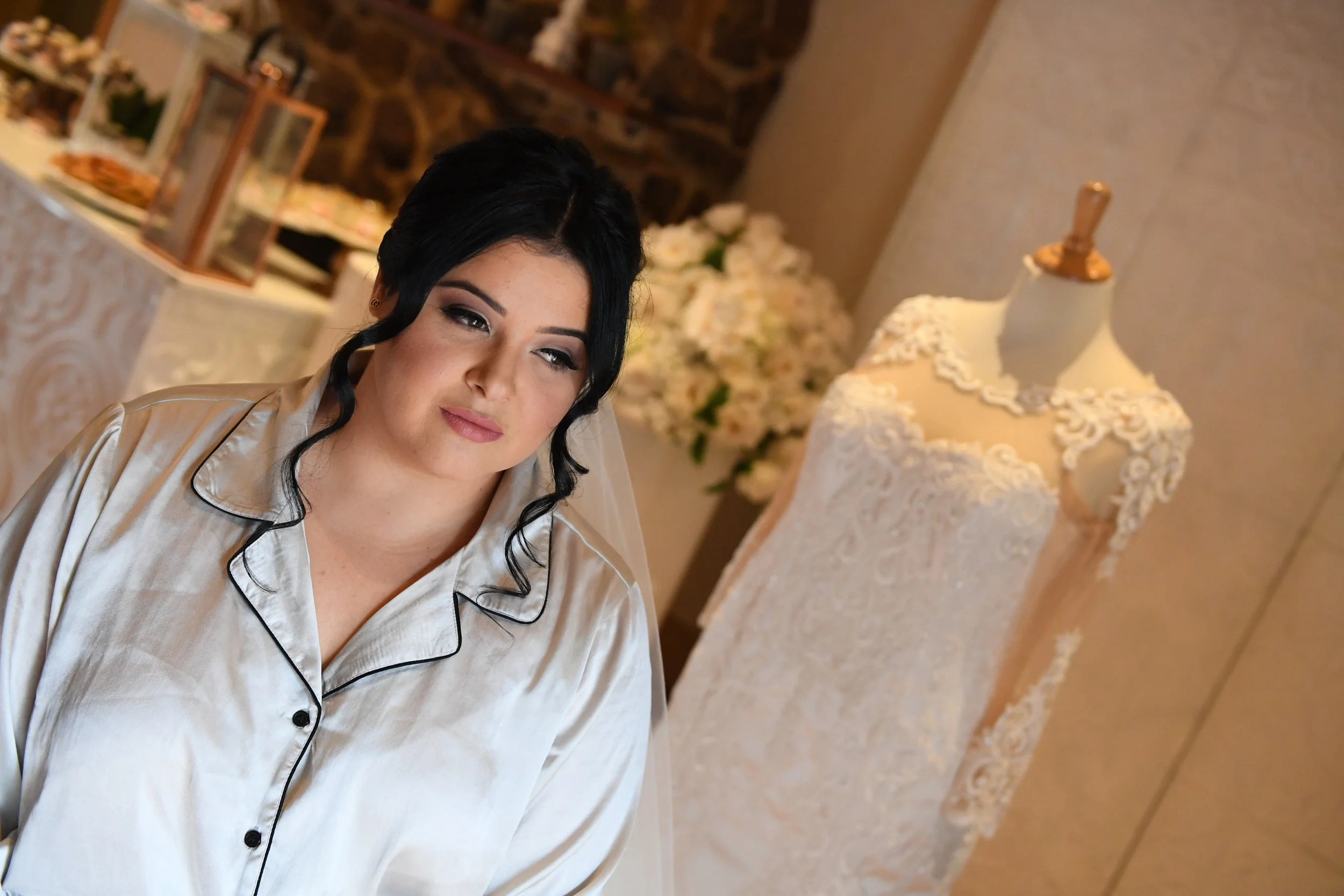 A woman with dark hair and makeup wearing satin pajamas, standing in a decorated room with a table set for a celebration, floral arrangements, framed photos, and a cake display.
