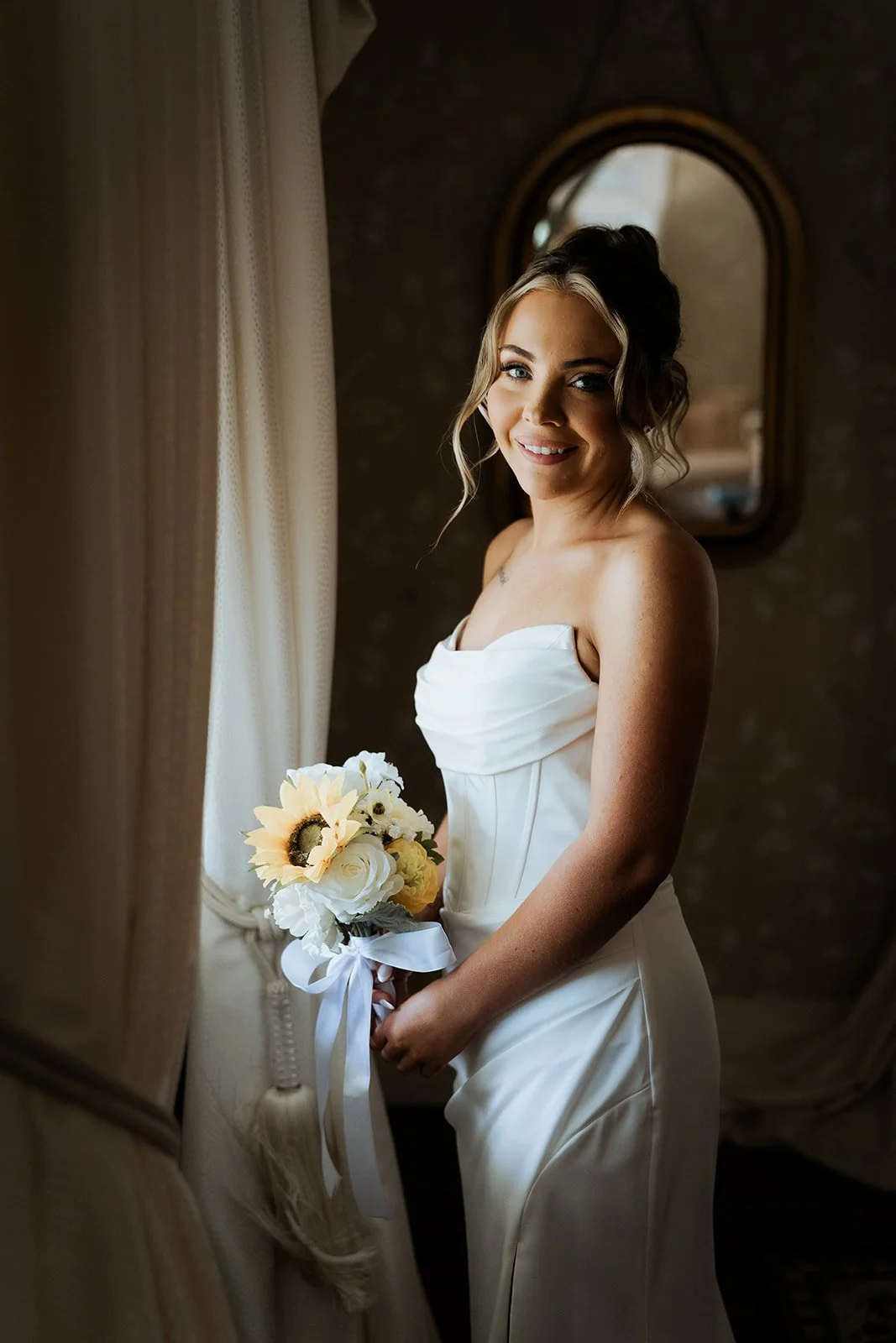 A smiling woman in a white wedding dress holding a bouquet of sunflowers and white roses, standing by a window with curtains, in front of a dark wall with a rounded mirror.