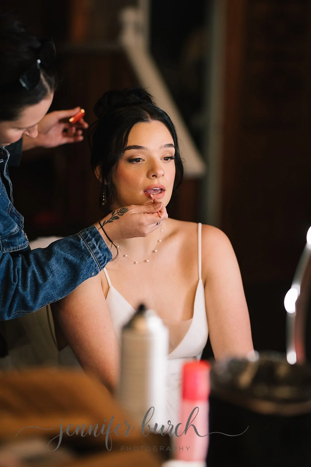 A woman with dark hair styled in an updo, wearing a white spaghetti strap dress and a pearl necklace, is getting her makeup done by a makeup artist. The makeup artist is applying makeup around her lips and is wearing a denim jacket.