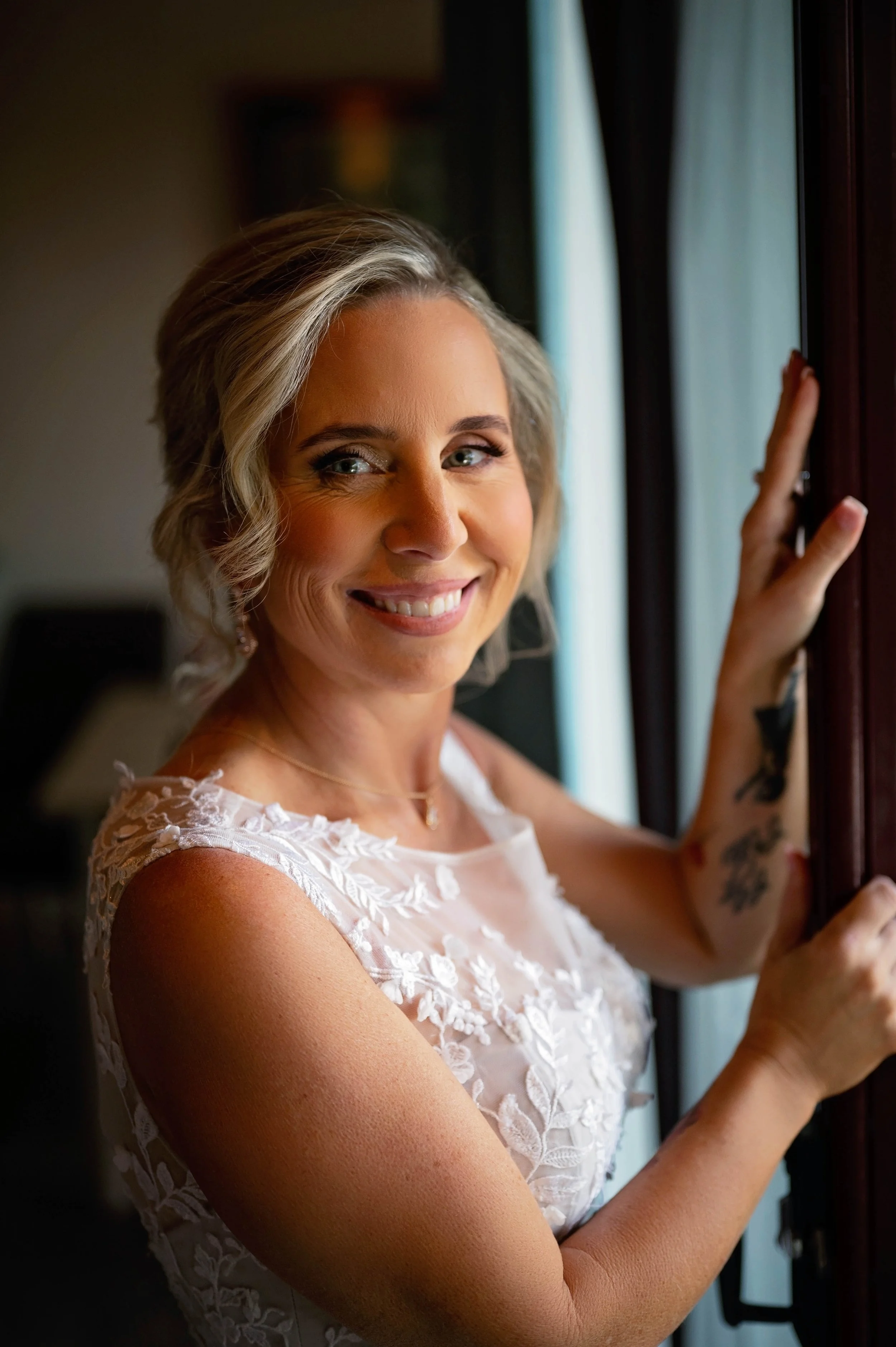 A smiling woman with blonde wavy hair wearing a white lace dress, touching a window frame with her right hand, standing in front of a curtain.