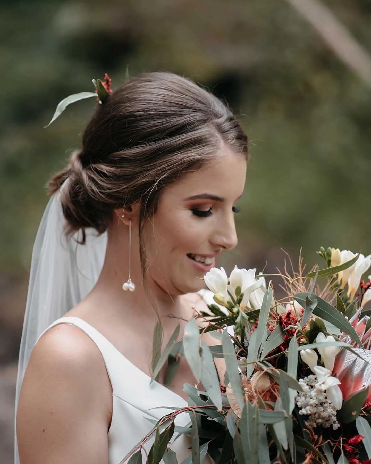 A smiling bride with brown hair styled in an elegant updo decorated with a small flower, wearing a white sleeveless wedding dress, holding a bouquet of white lilies, roses, and greenery, outdoors with blurred green foliage in the background.