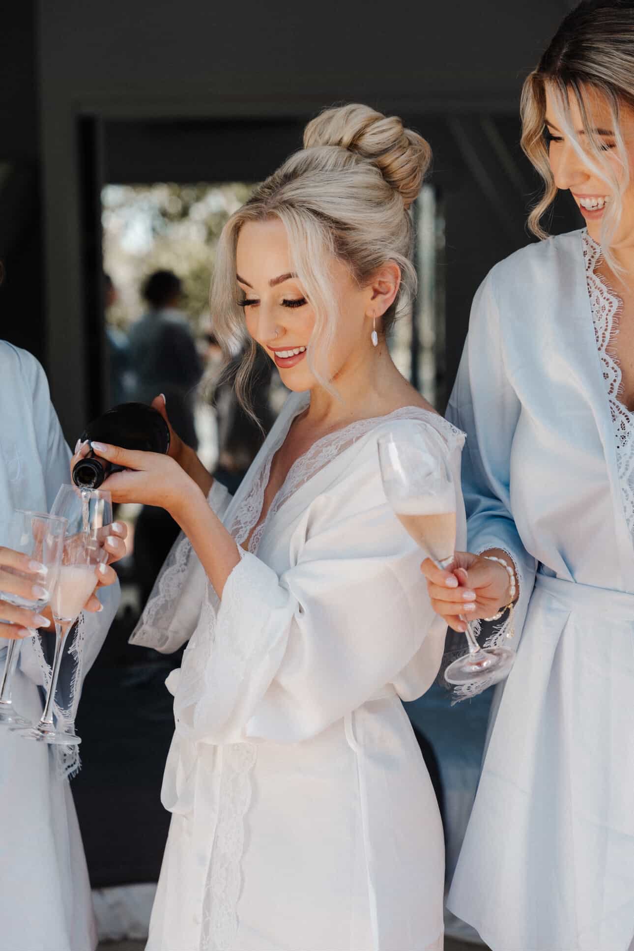 Women in white robes celebrating with champagne at an outdoor event, one woman pouring wine into a glass.