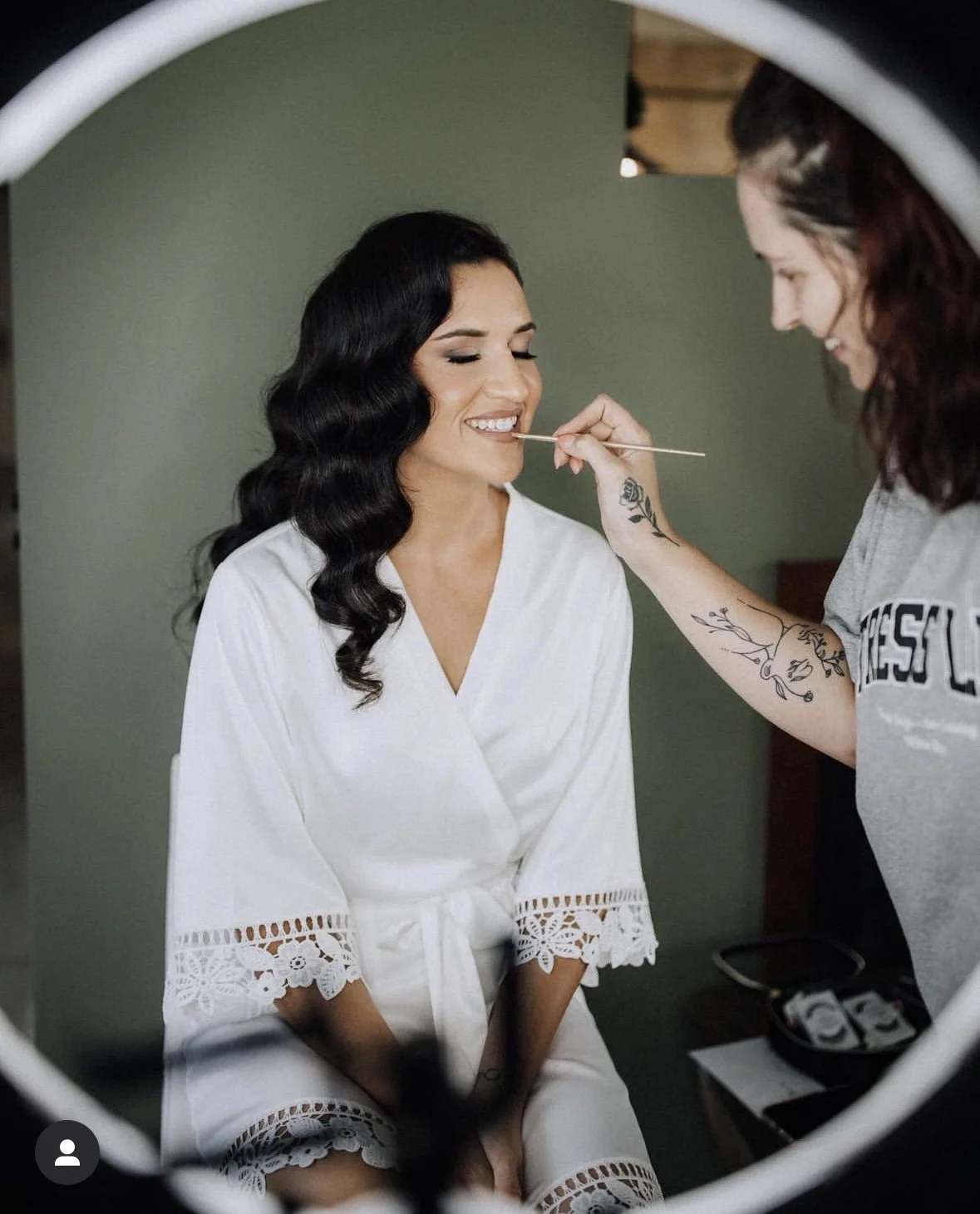 A woman with long, wavy dark hair sitting on a chair, getting her makeup done by a makeup artist with tattoos on her arm. The woman is smiling with her eyes closed, wearing a white robe with lace details on the sleeves and hem.