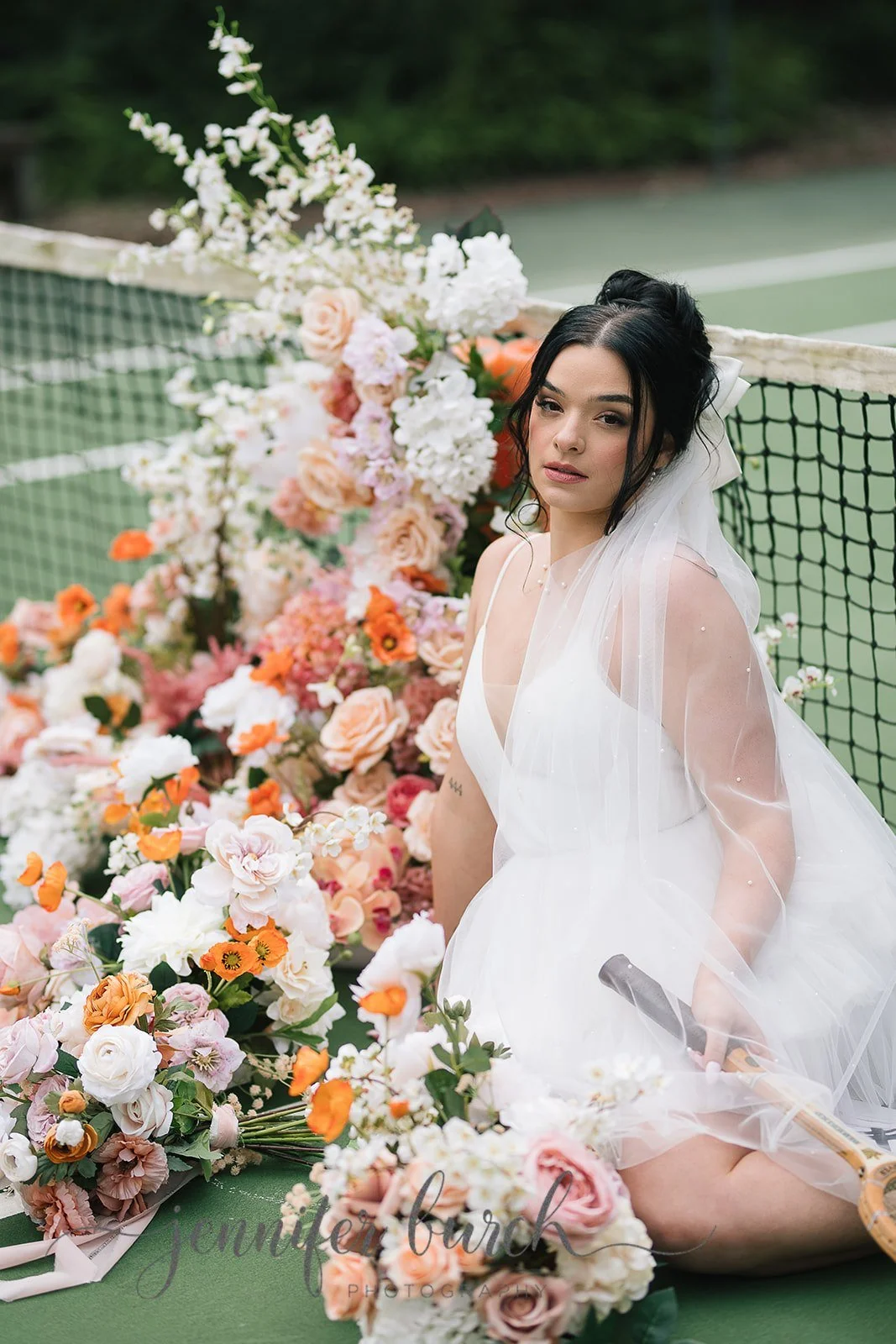 A bride with dark hair in a white dress and veil pose on a tennis court surrounded by flowers.