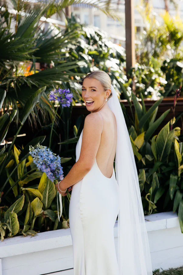 Smiling bride in a white backless wedding dress holding a bouquet of blue flowers, standing outdoors among green plants.