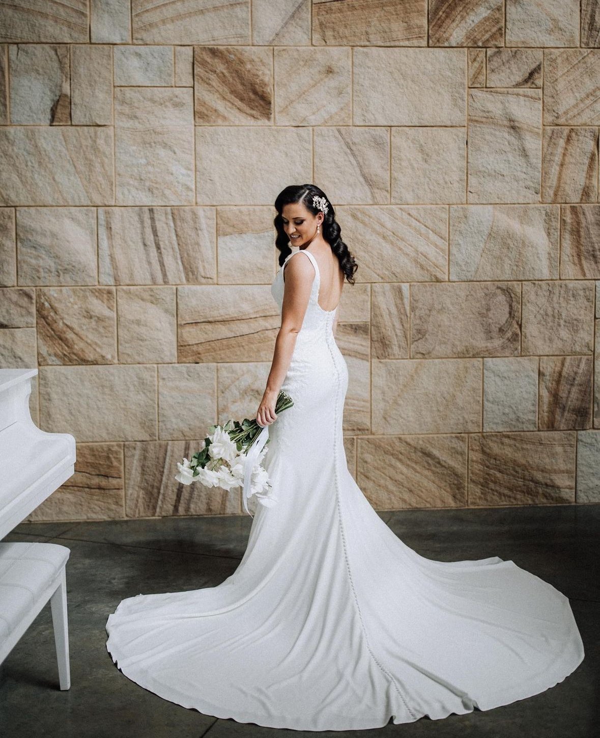 A bride in a white wedding gown with a long train, holding a bouquet of white lilies, standing against a textured stone wall.
