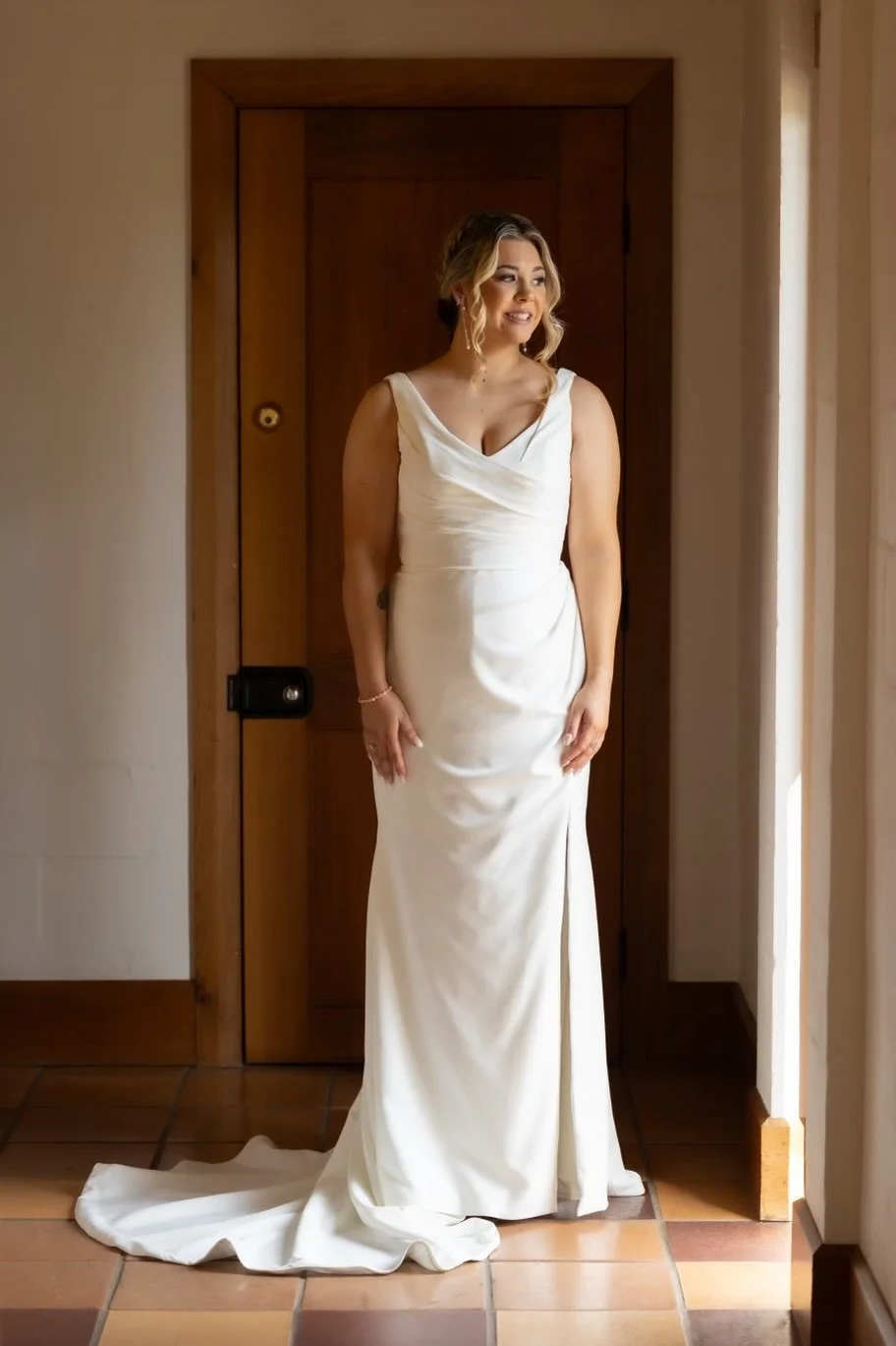 A woman in a white wedding dress standing indoors near a wooden door, smiling and looking away.