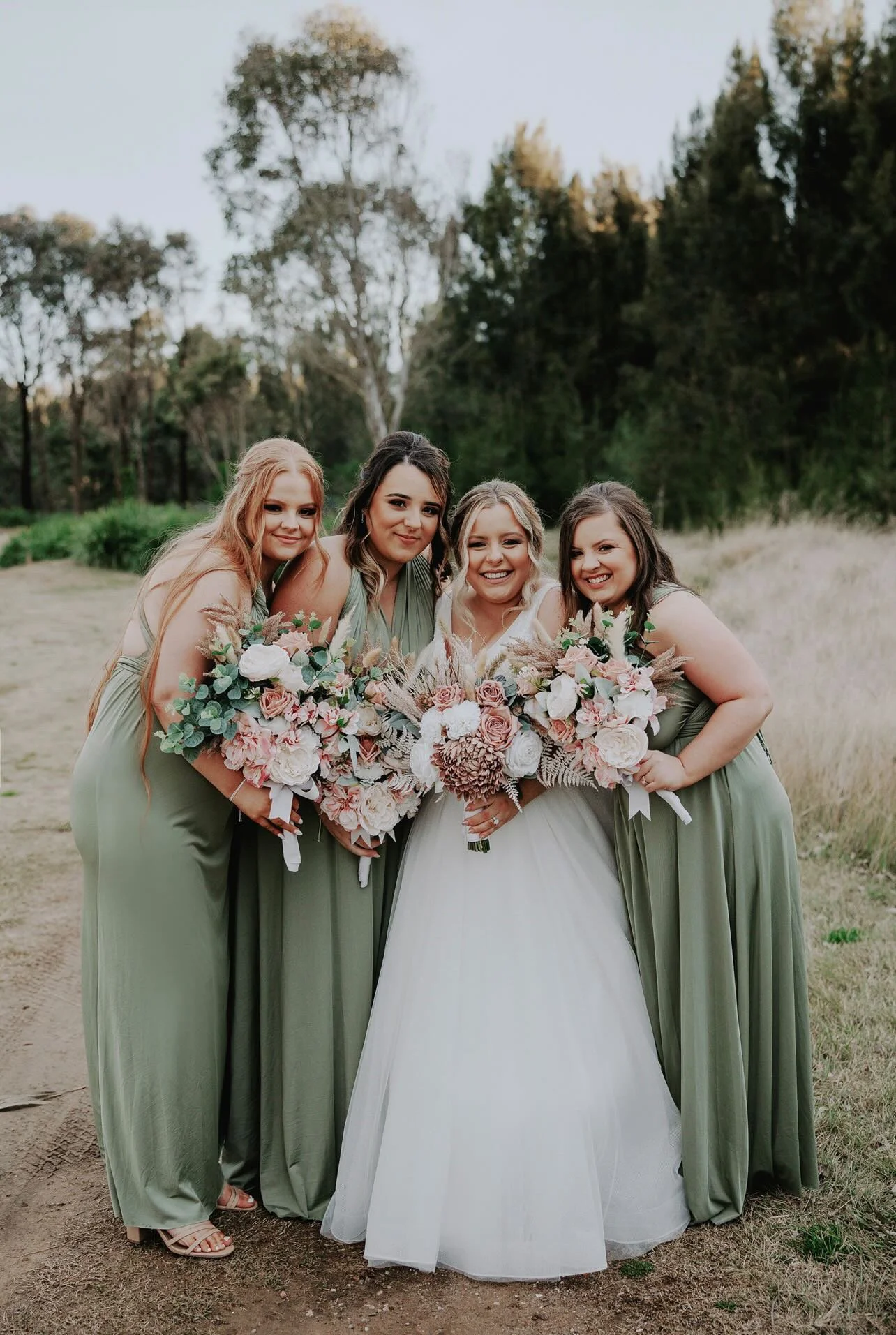 Four women in green dresses at an outdoor wedding, holding large bouquets of pink, white, and purple flowers, standing on a dirt path with trees and open field in the background.