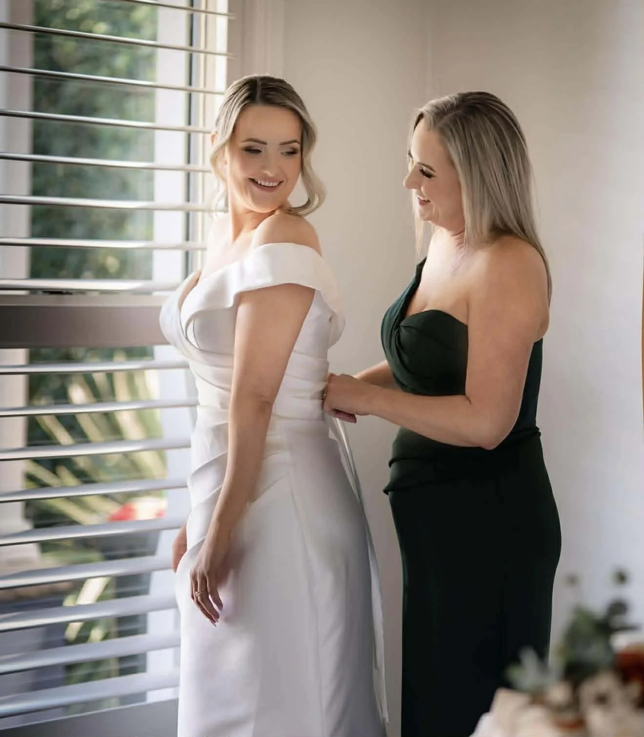 A bride in a white wedding dress smiling at a woman in a black dress who is helping her with her gown near a window with horizontal blinds.