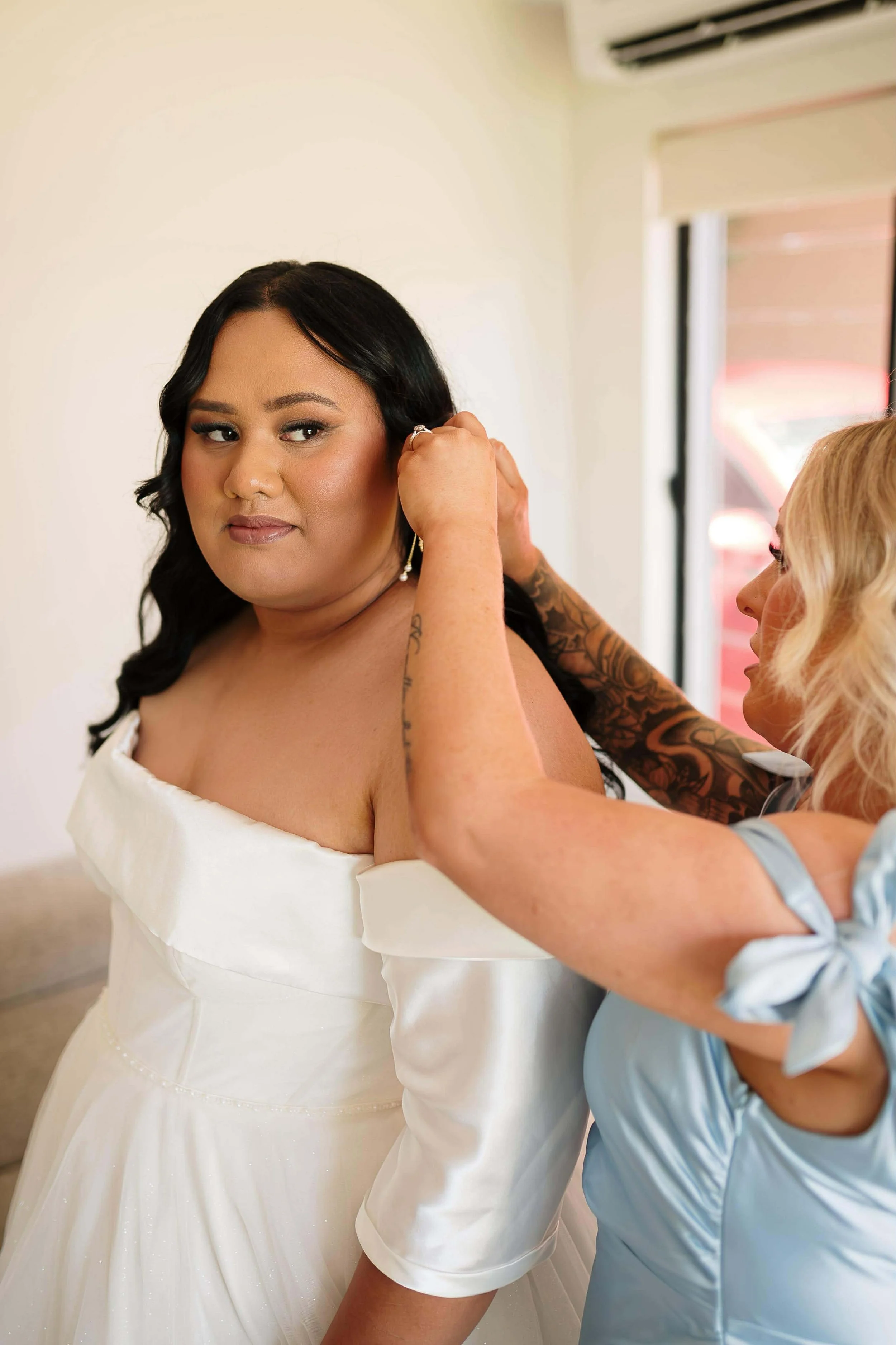 A bride in a white dress getting dressed by a helper, who is in a light blue shirt, in a room with a window and an air conditioner.