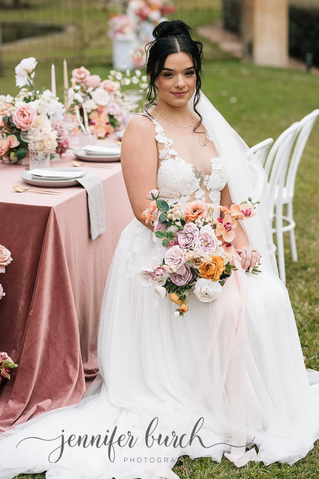 A bride in a white wedding dress holding a bouquet of pink, purple, white, and orange flowers, sitting outdoors with a decorated table of flowers and candles in the background.