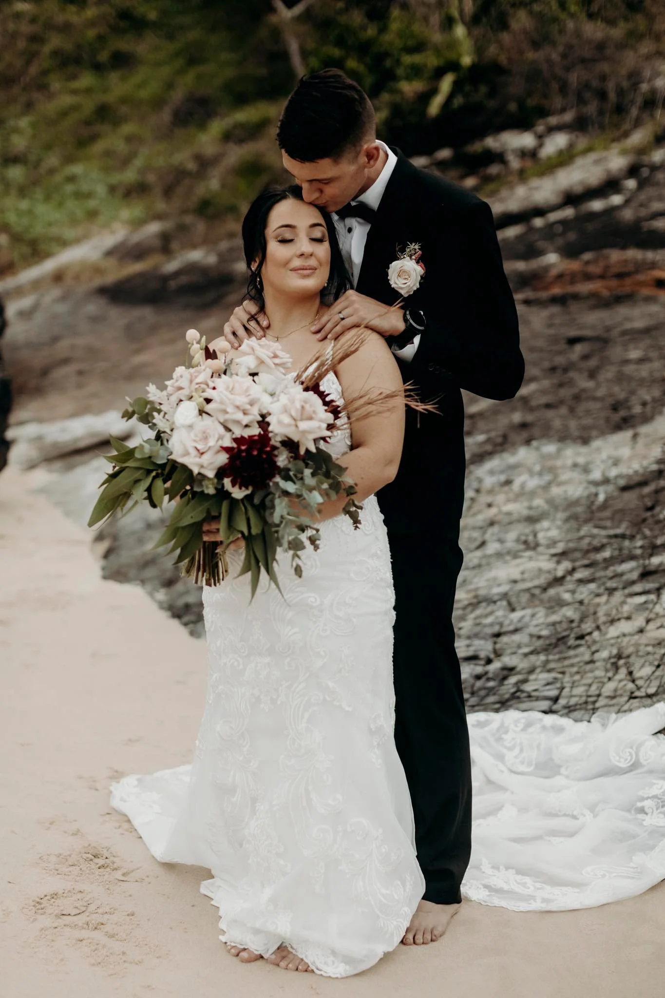 A bride and groom standing together on a beach, with the groom kissing the bride on her forehead. The bride is holding a large bouquet of flowers and is wearing a white lace wedding dress. The groom is wearing a black tuxedo with a boutonniere. They 
