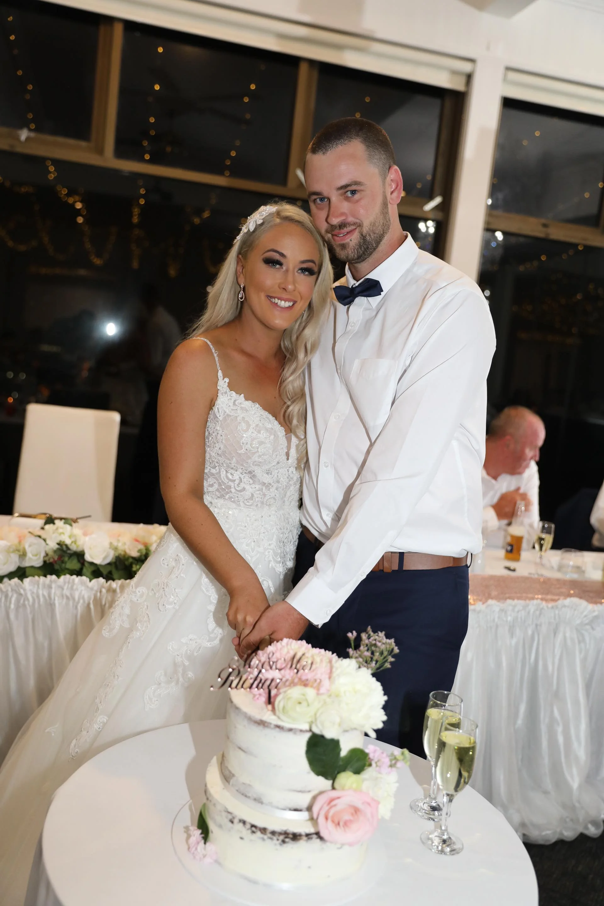 A newlywed couple smiling and holding hands during their wedding reception, with a wedding cake adorned with flowers on a table in front of them.