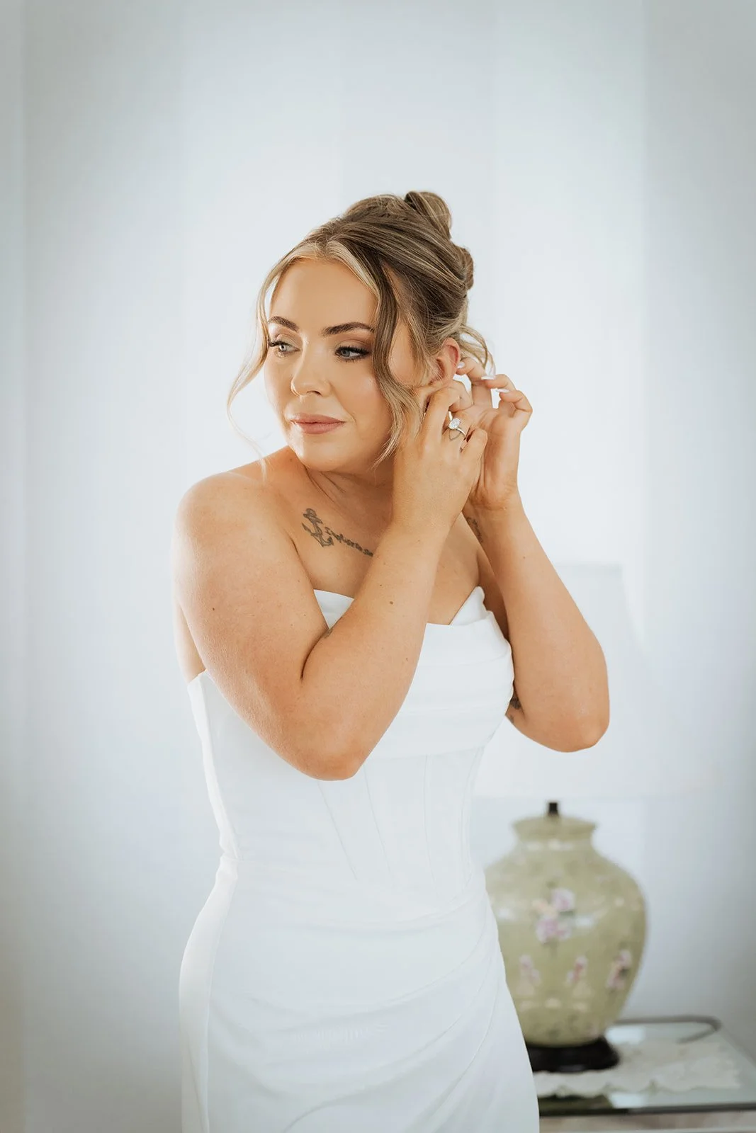 A woman in a white strapless dress is putting on an earring, with elegant makeup and styled hair, standing in a bright room with a vase in the background.