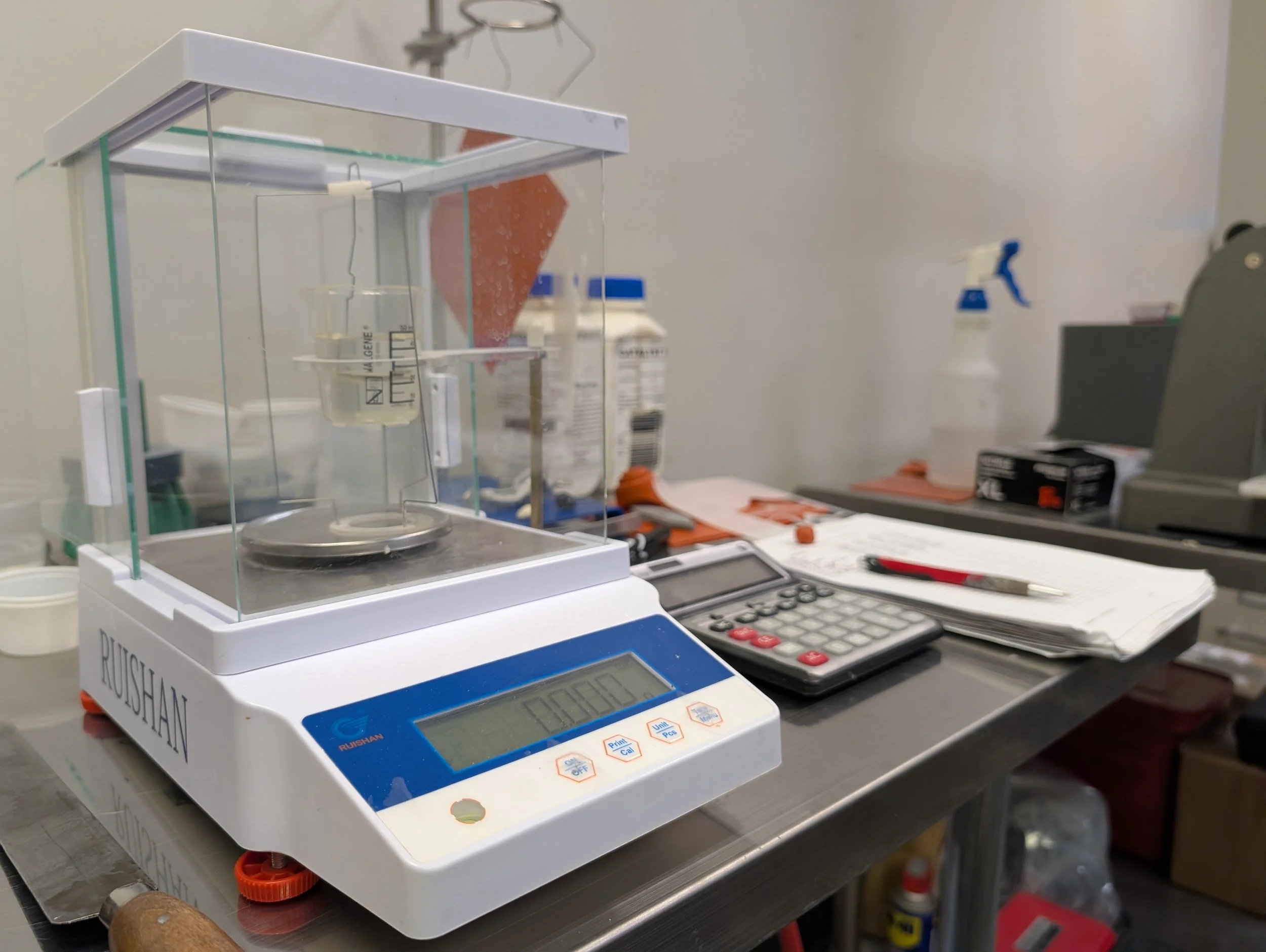 Laboratory countertop with a digital scale in the foreground, scientific supplies, bottles, a computer monitor, and various tools in the background.