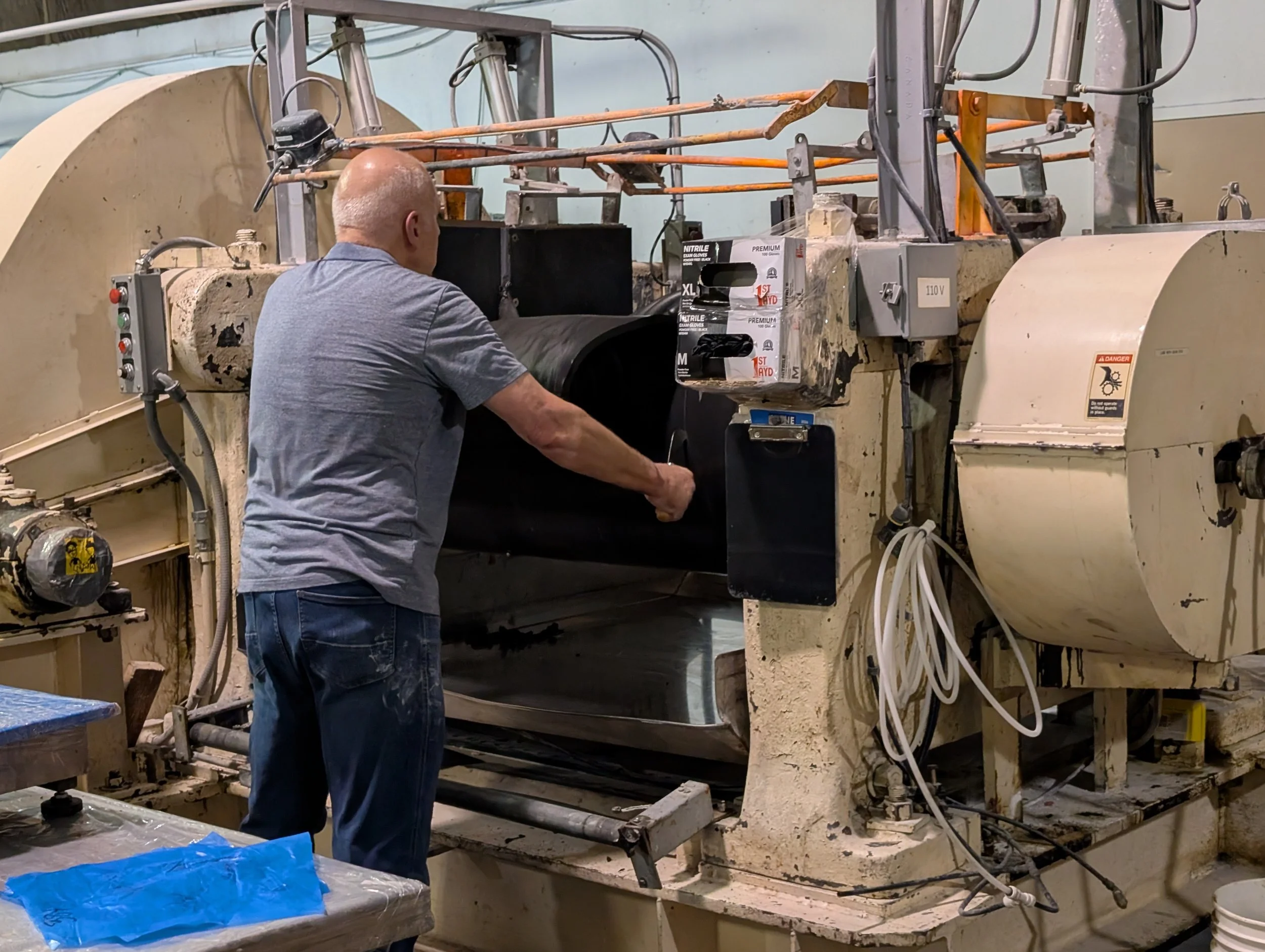 A man with a shaved head and gray t-shirt working with a large industrial machine in a workshop or factory. The machine is dirty and appears to be used for manufacturing or processing, with various pipes, wires, and controls visible.
