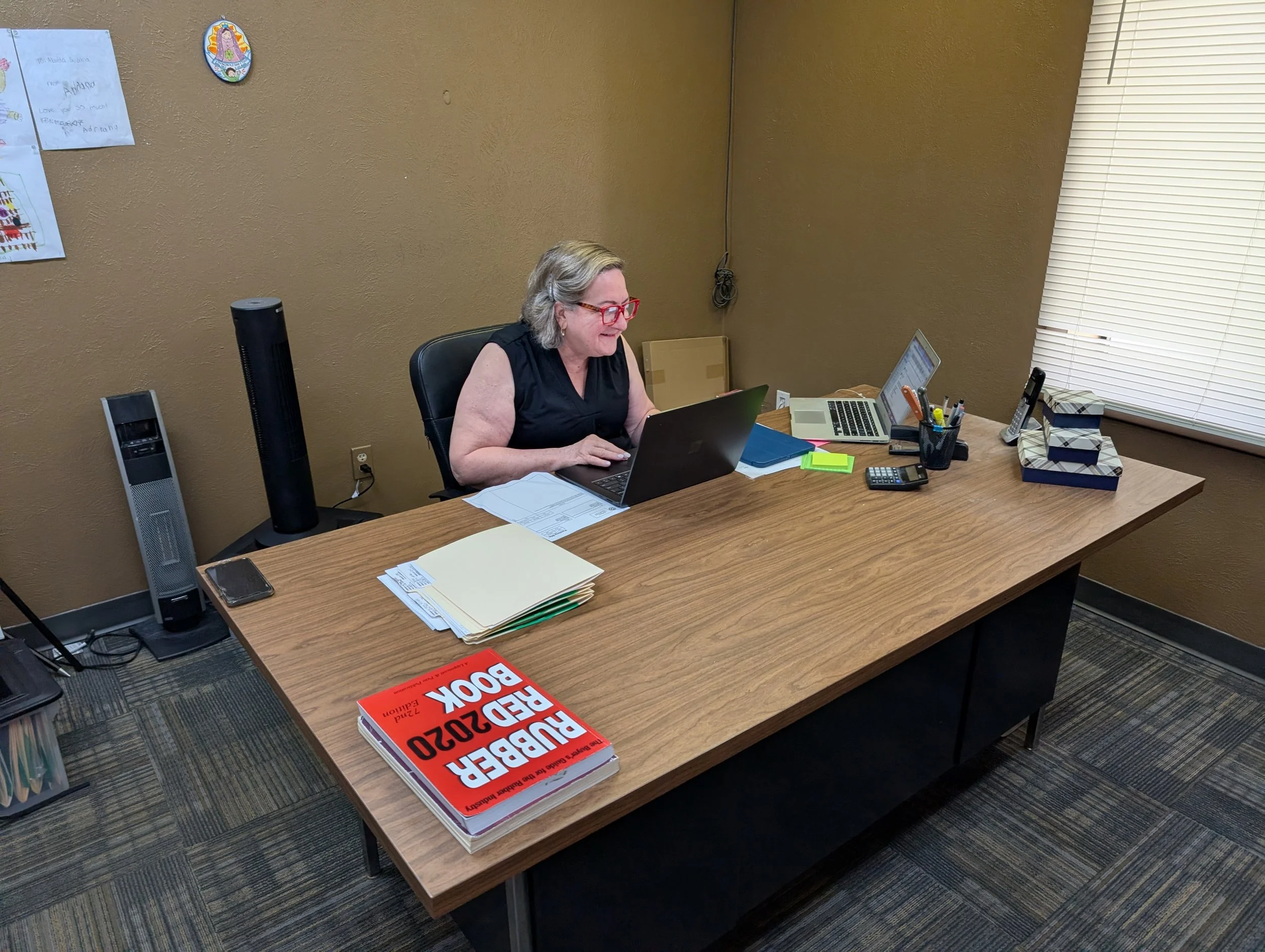 An office room with a woman wearing glasses, working on a laptop, seated at a wooden desk with books, papers, a phone, a walkie-talkie, and office supplies. A tall fan is beside her, with a wall decorated with a religious picture and drawings, and a heat vent at the bottom.