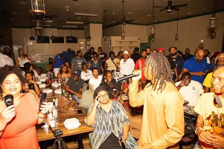 A lively indoor Israelites gathering with a speaker addressing the audience, who are seated at tables and standing, enjoying drinks and taking photos. New Legacy Initiative. New Legacy, Inc.