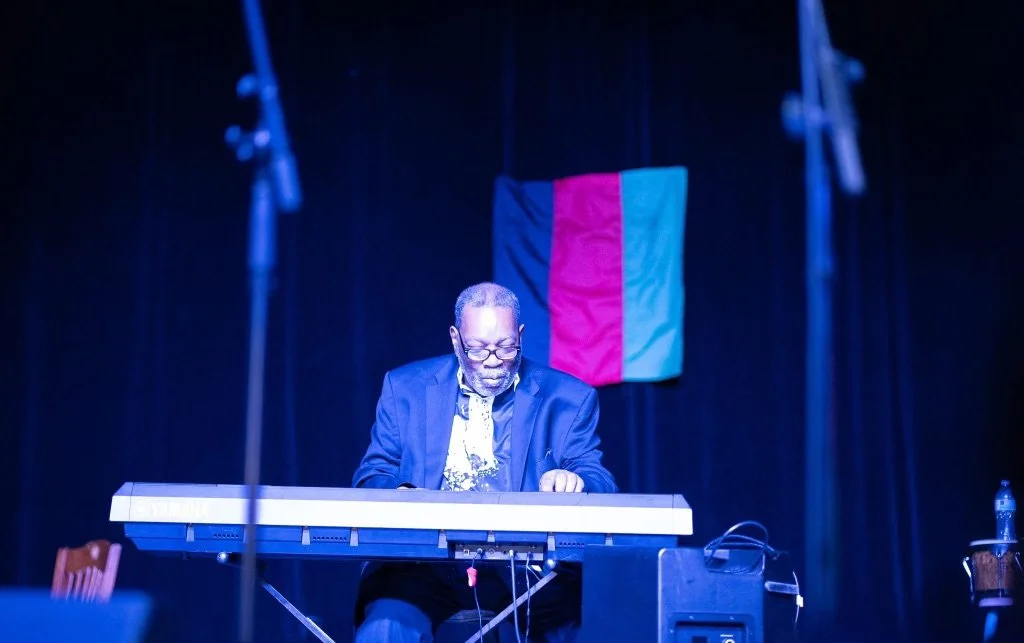 An Israelite man playing keyboard on stage with a black curtain backdrop and a pink and blue flag hanging behind him. New Legacy Initiative. New Legacy, Inc.